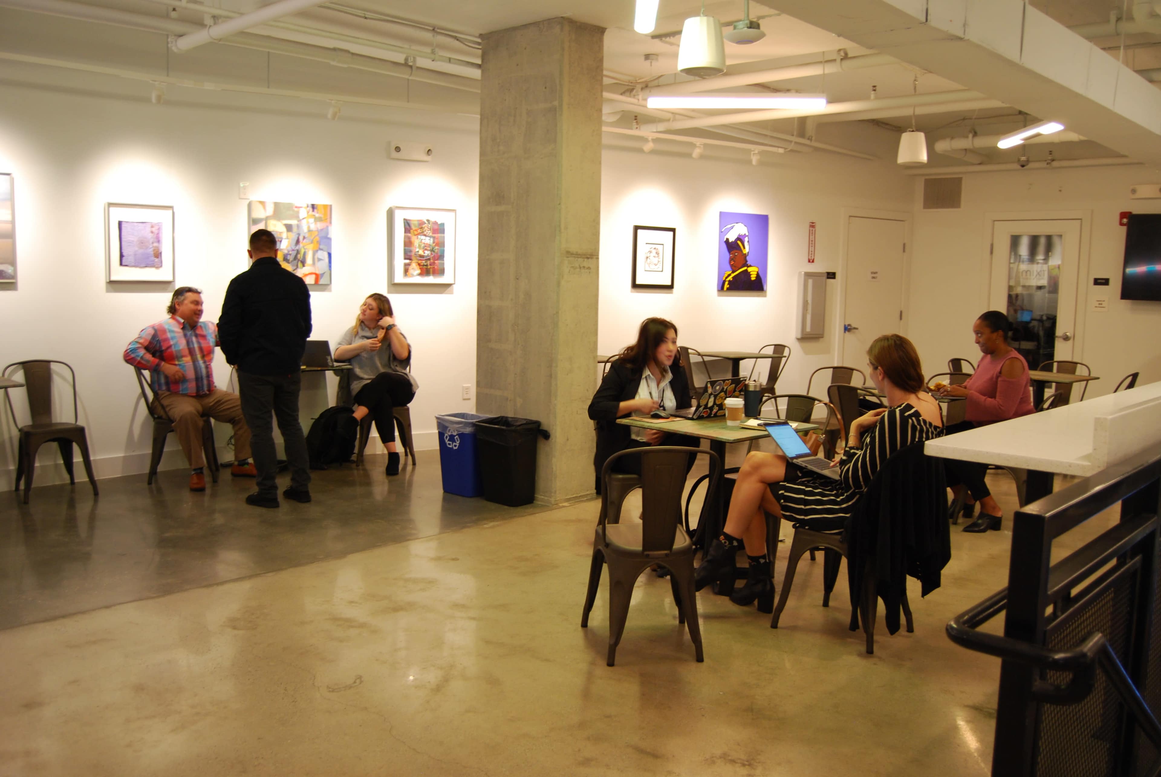 A group of people works and interacts in a modern workspace that features metal chairs and artwork on the walls.