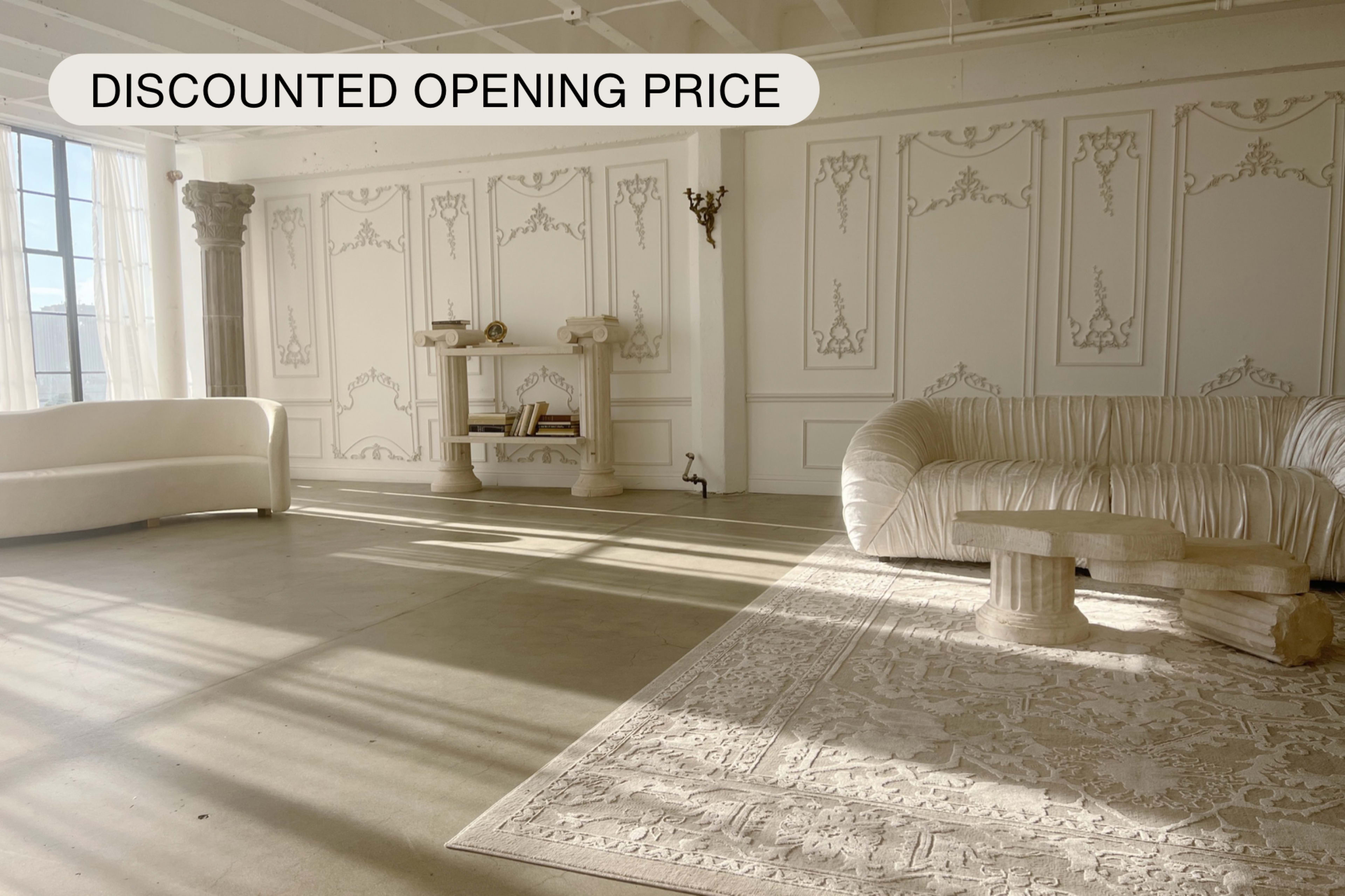 A spacious room features a textured white sofa, a stone-like coffee table, and a shelf against a backdrop of ornate wall paneling.