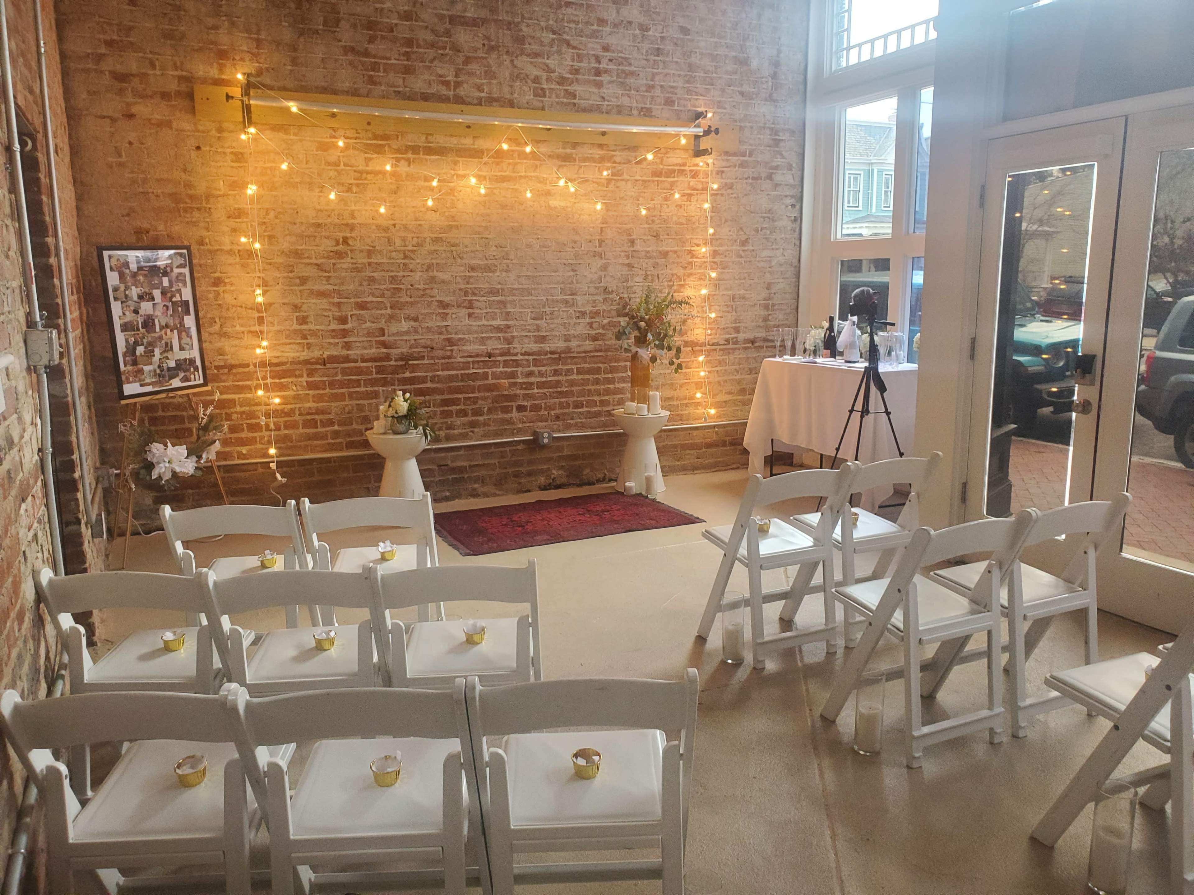 A small wedding ceremony setting features white folding chairs arranged in front of a decorated backdrop with string lights, flanked by a table and floral arrangements.
