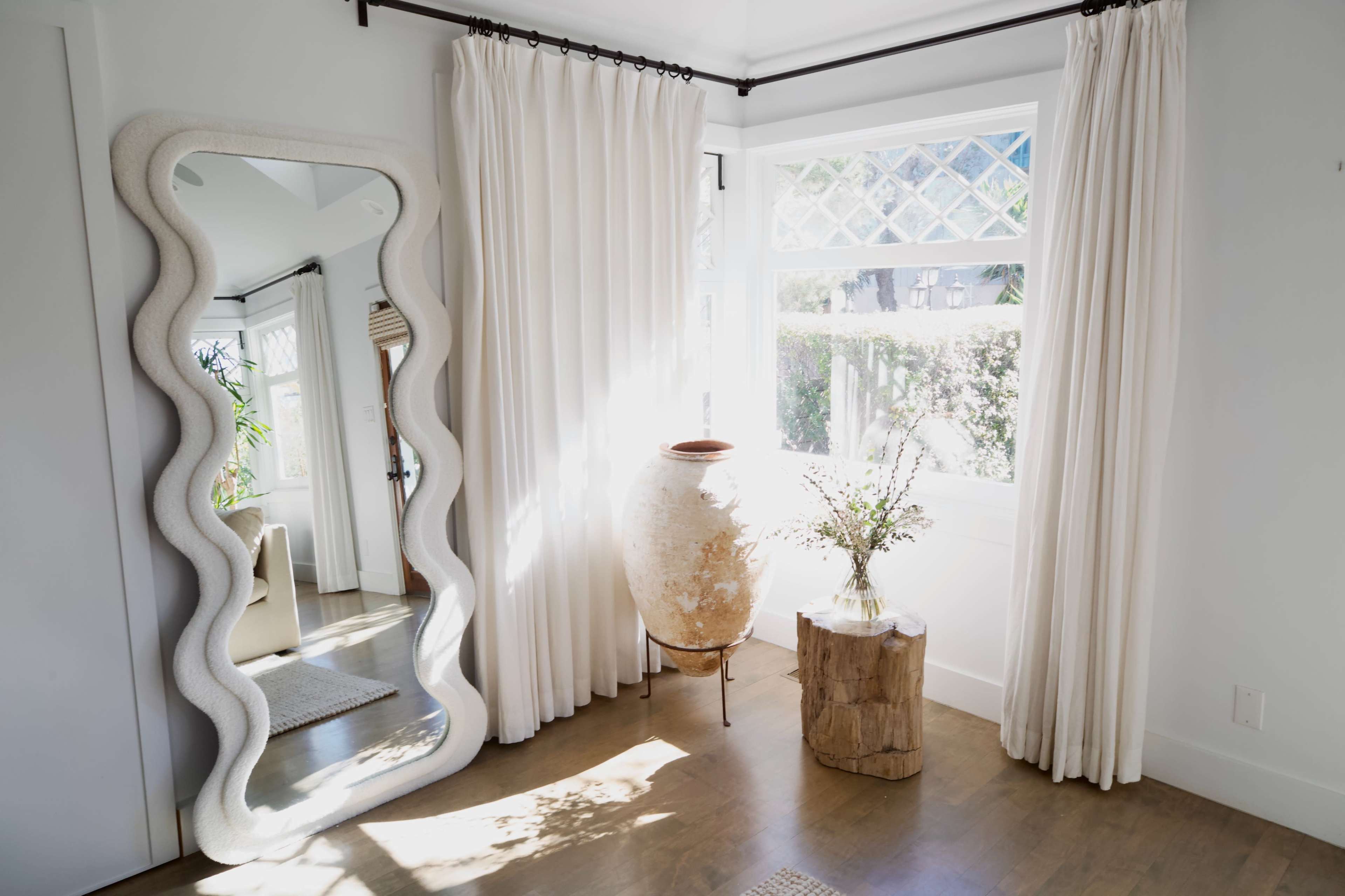 A large, wavy-edged mirror leans against a white wall next to a window with sheer curtains and a decorative pot on a wooden side table.