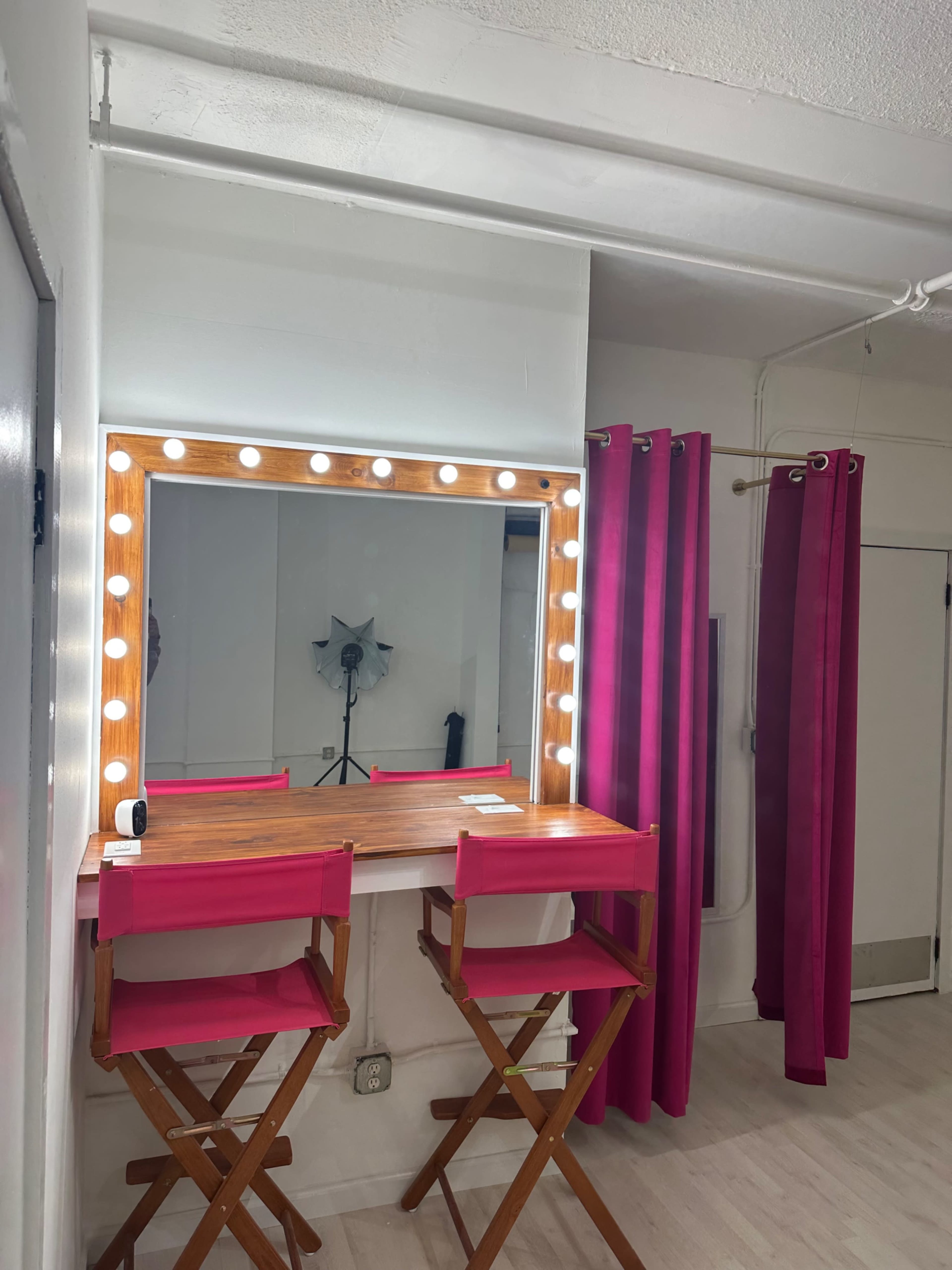 The scene features a well-lit makeup vanity with a large mirror surrounded by lights, two pink director's chairs, and a backdrop curtain in a room with light-colored walls and flooring.