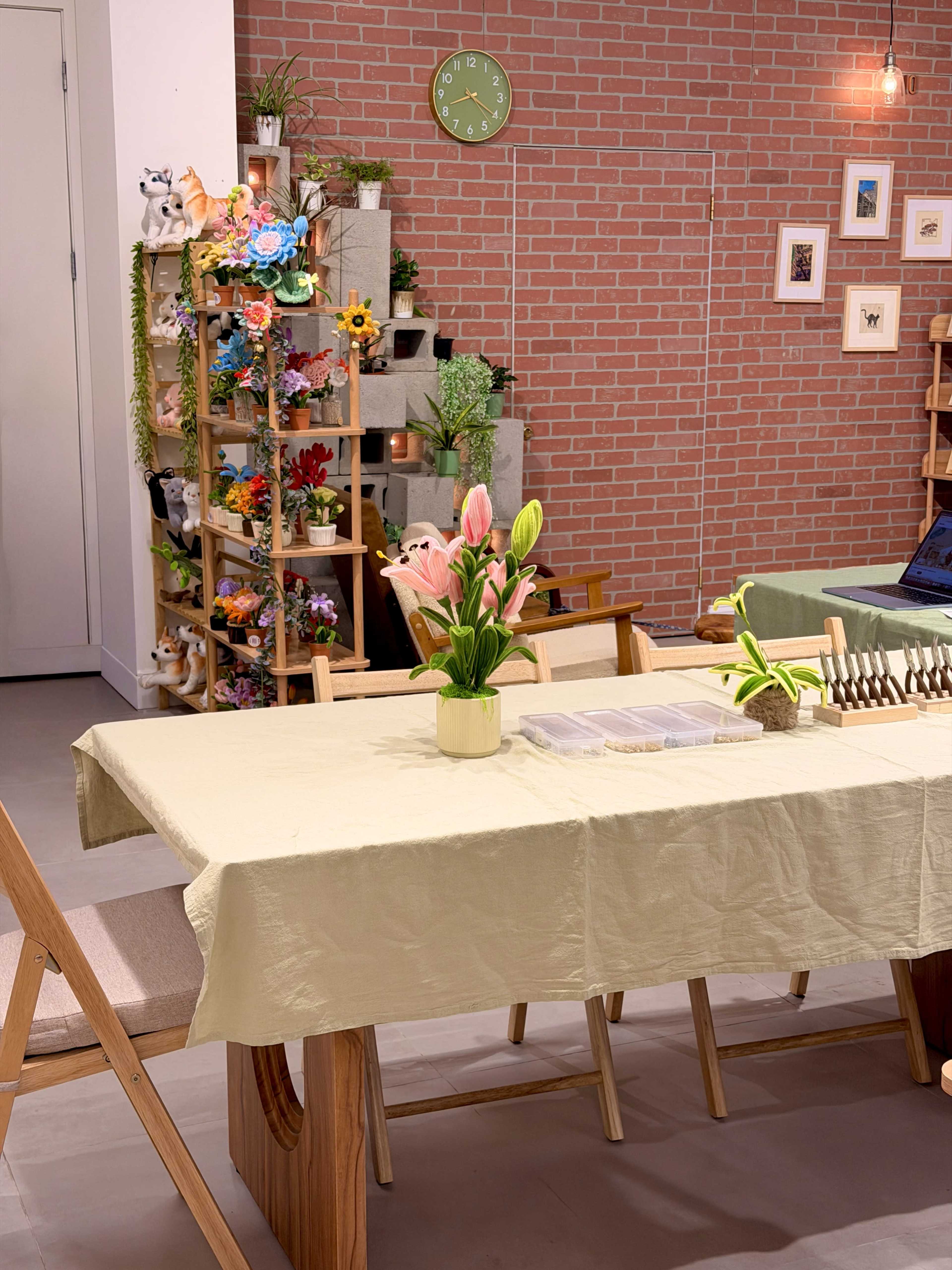 The image shows a cozy dining area with a light-colored tablecloth, a vase of flowers on the table, and decorative plants and artwork on the brick wall behind.