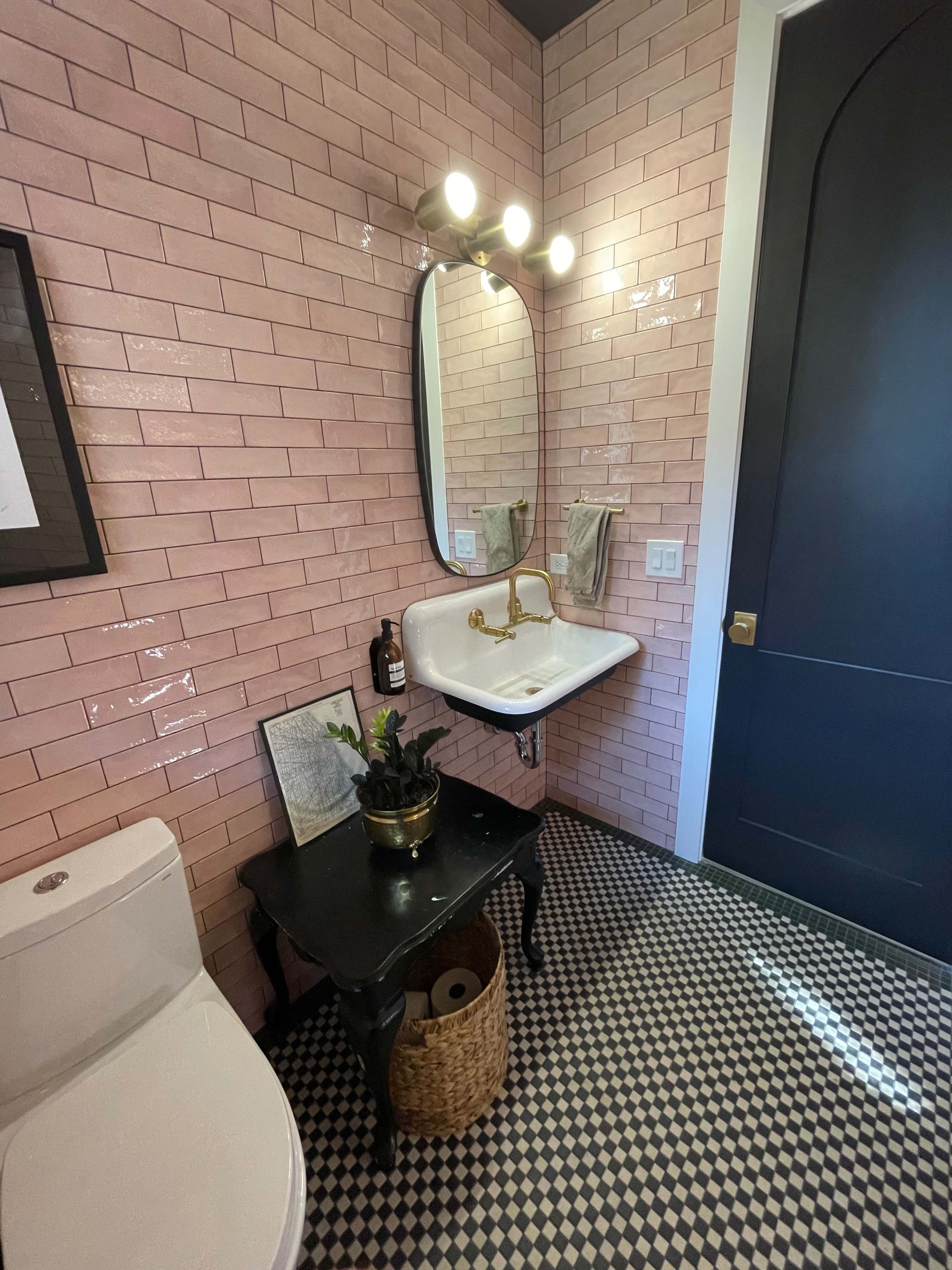 A small bathroom with pink tiled walls, a white sink mounted on a black table, a round mirror, and a black door, complemented by a checkered floor.