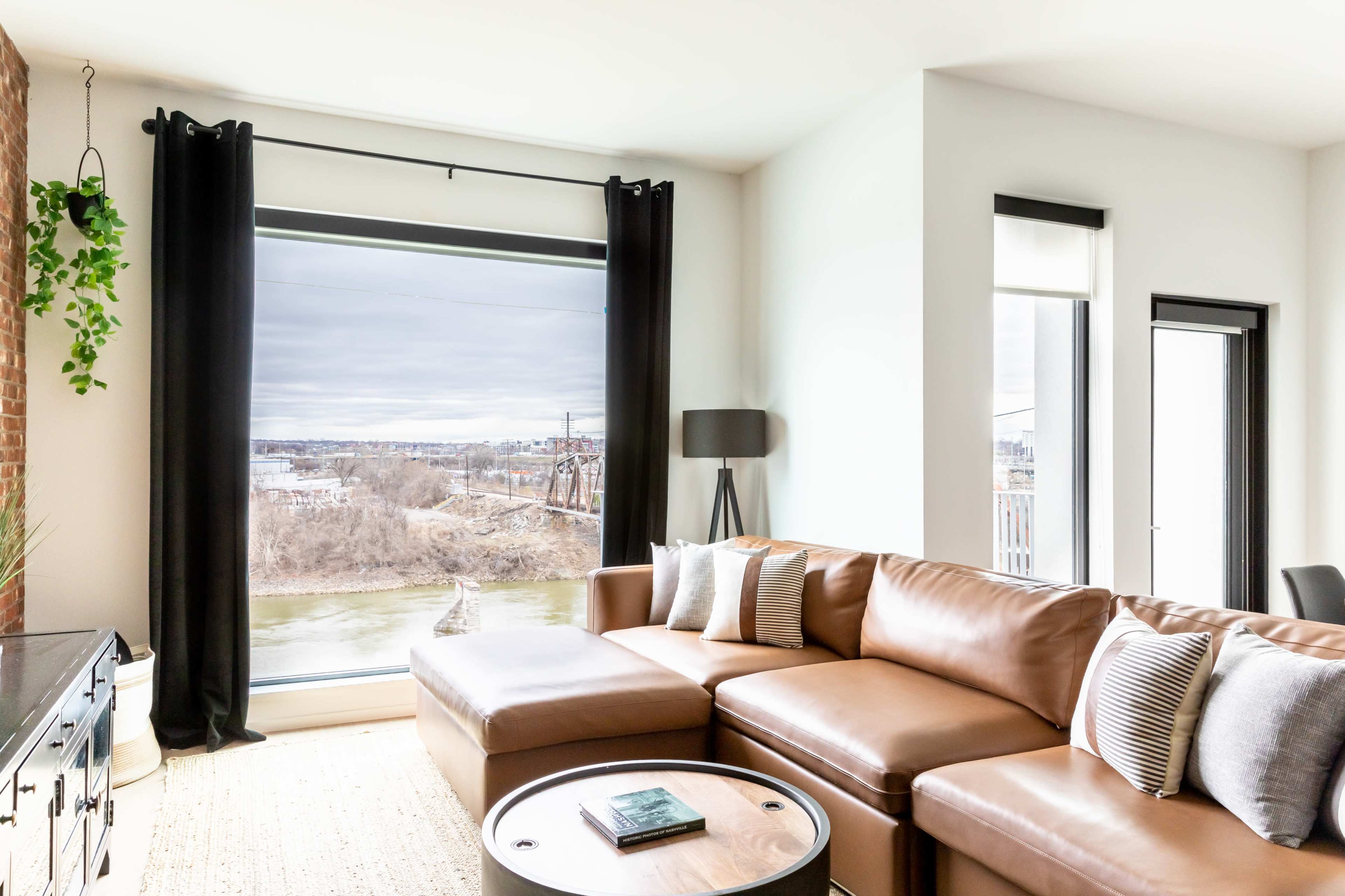A modern living room features a large window overlooking a river, with a brown sectional sofa and a round coffee table.