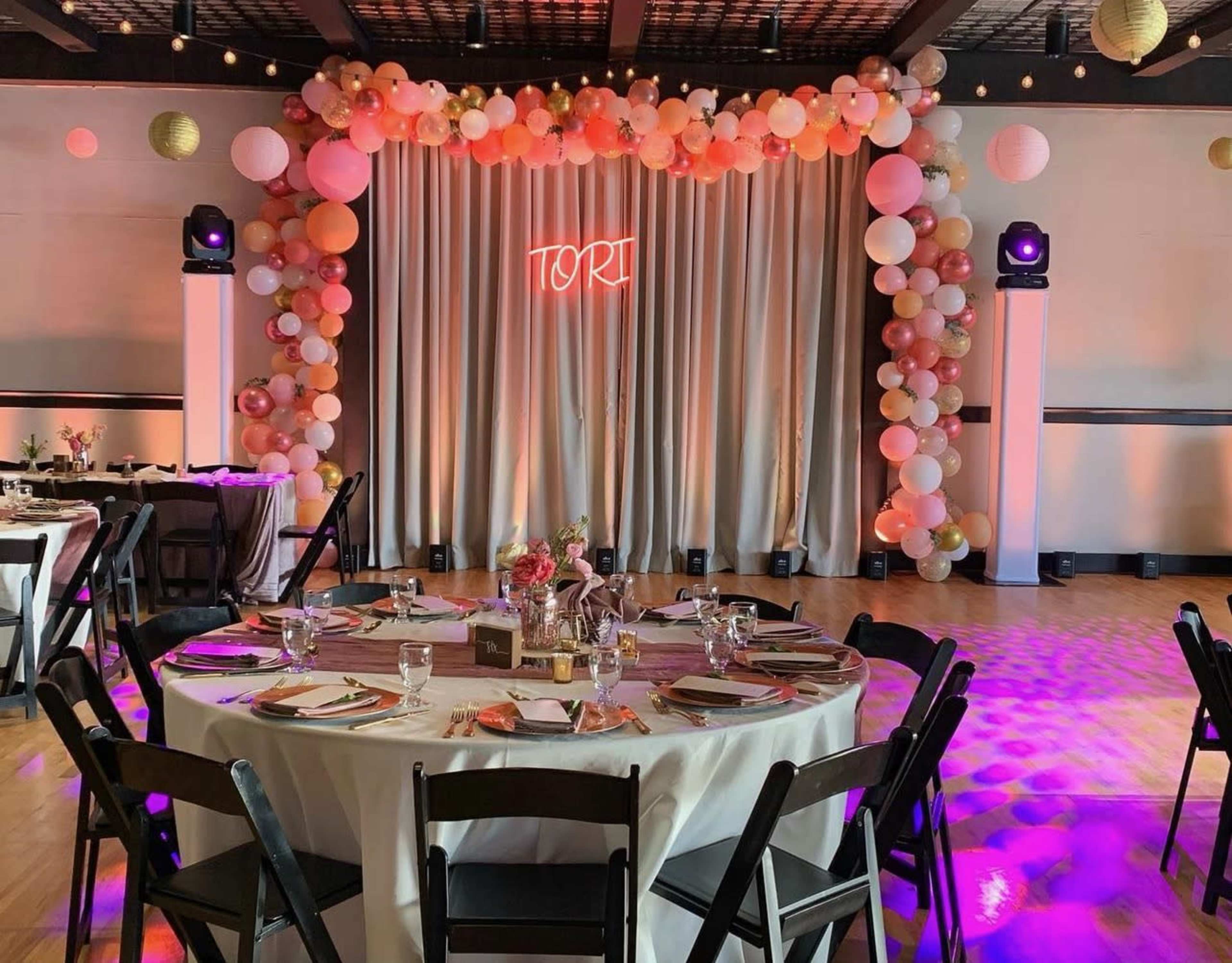 A decorated event space with a table set for dining, featuring a backdrop of balloons and a glowing sign that reads "Tori."