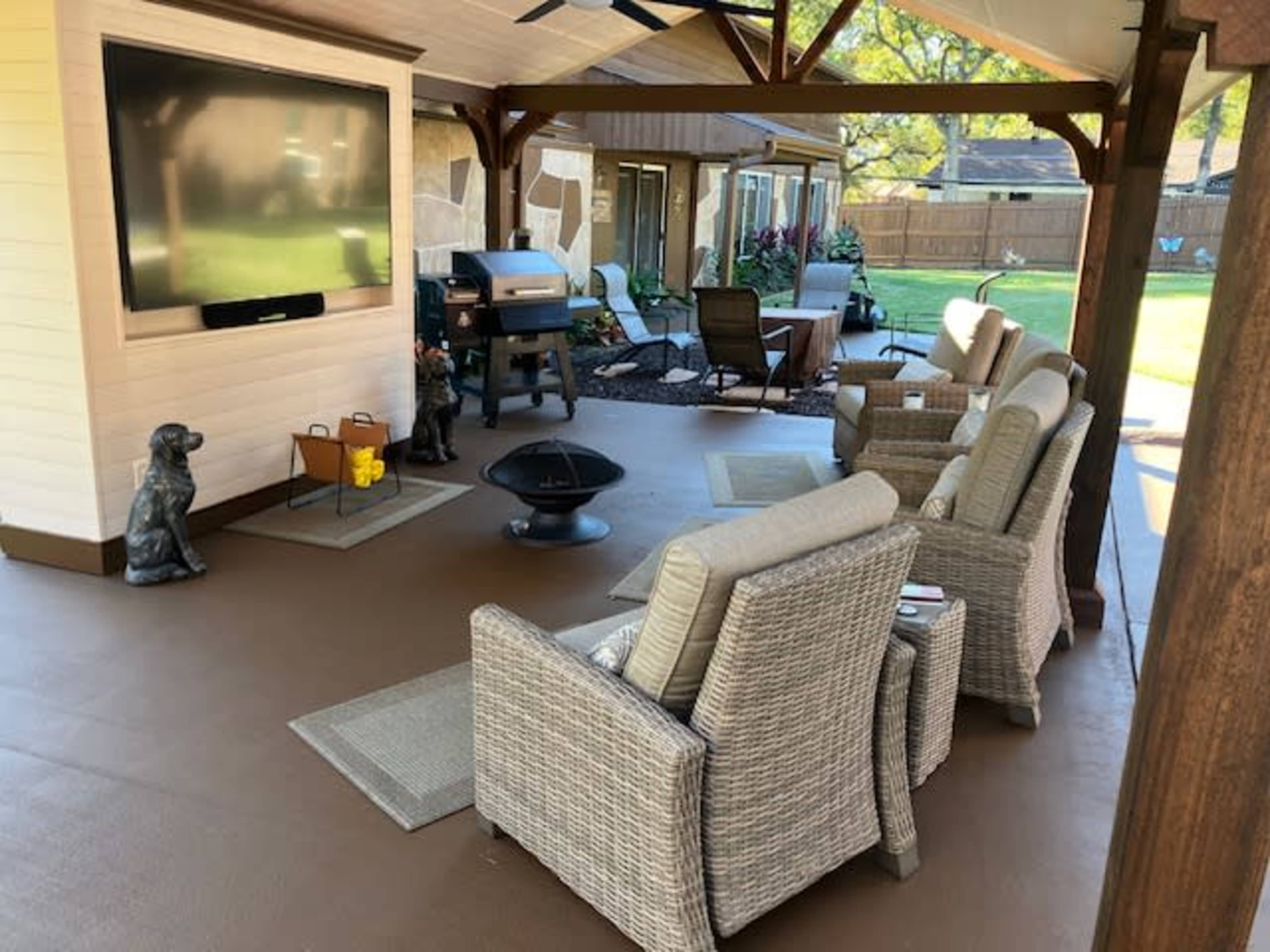 The image shows a covered patio area featuring wicker chairs, a fire pit, a television mounted on the wall, and a grill in the background.