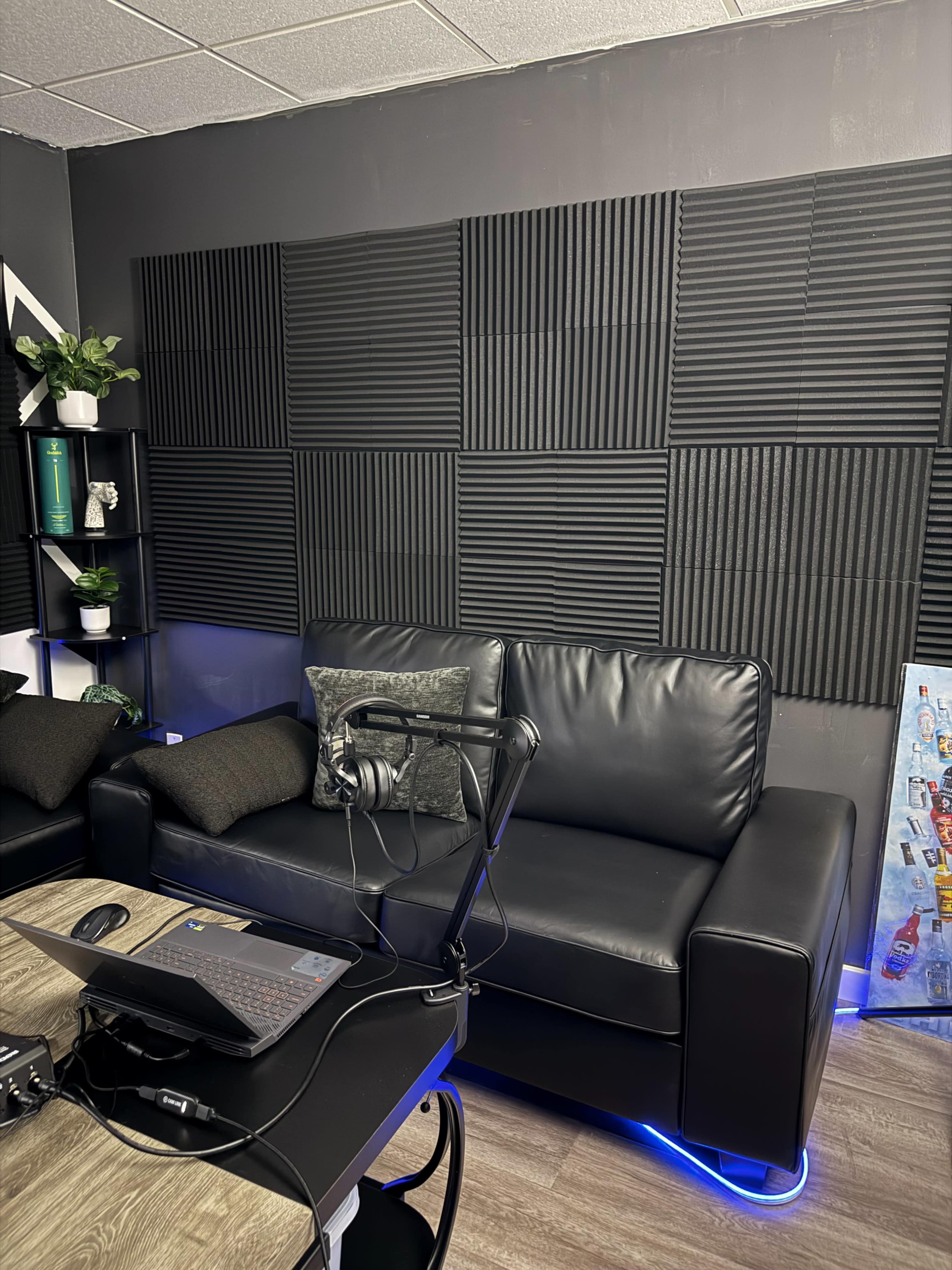The image shows a modern studio space with a black leather couch, acoustic foam panels on the wall, a laptop on a desk, and a microphone setup.