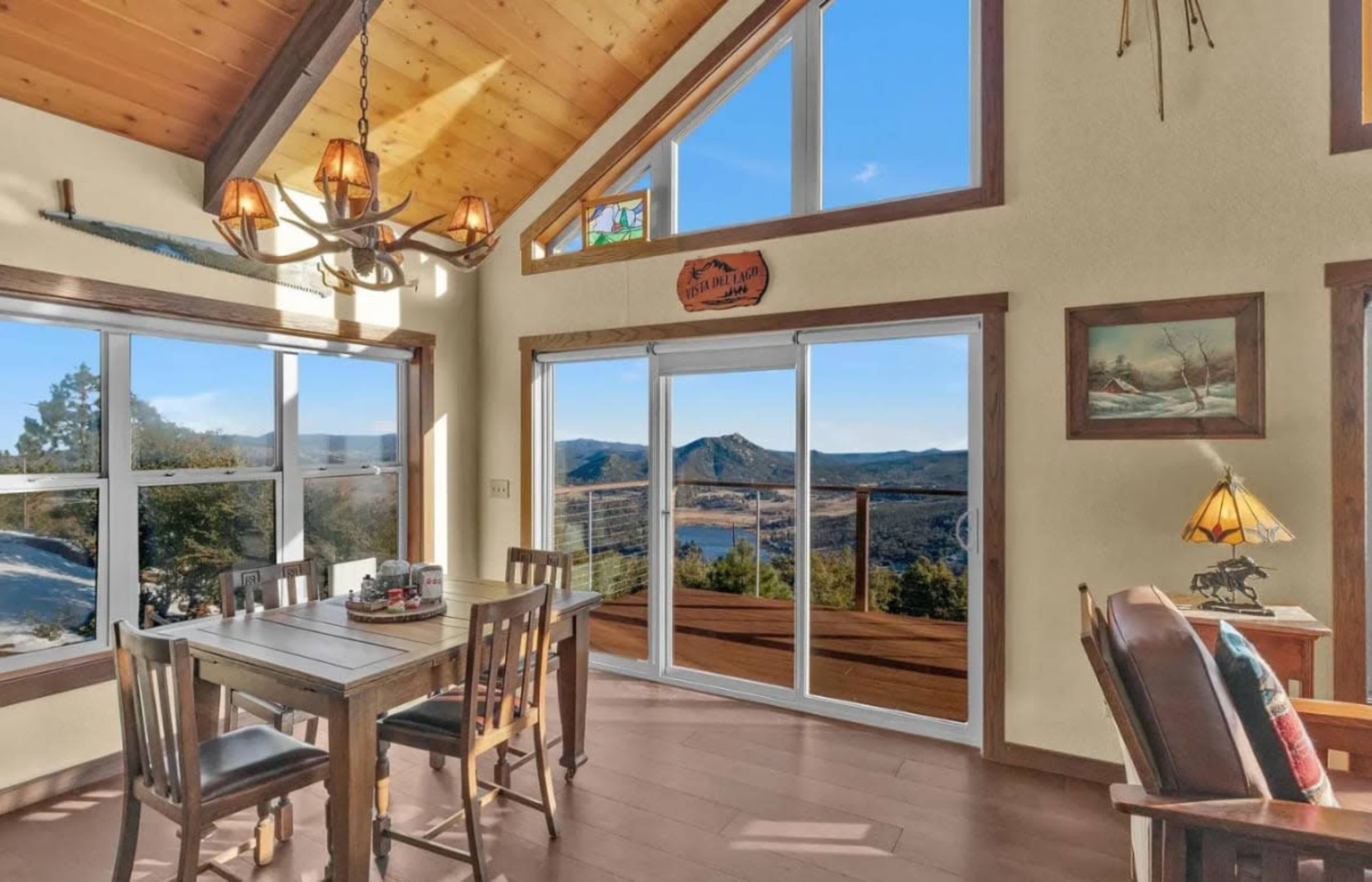 The image shows a rustic dining area with a wooden table and chairs, large windows revealing a mountainous landscape, and a vaulted ceiling.