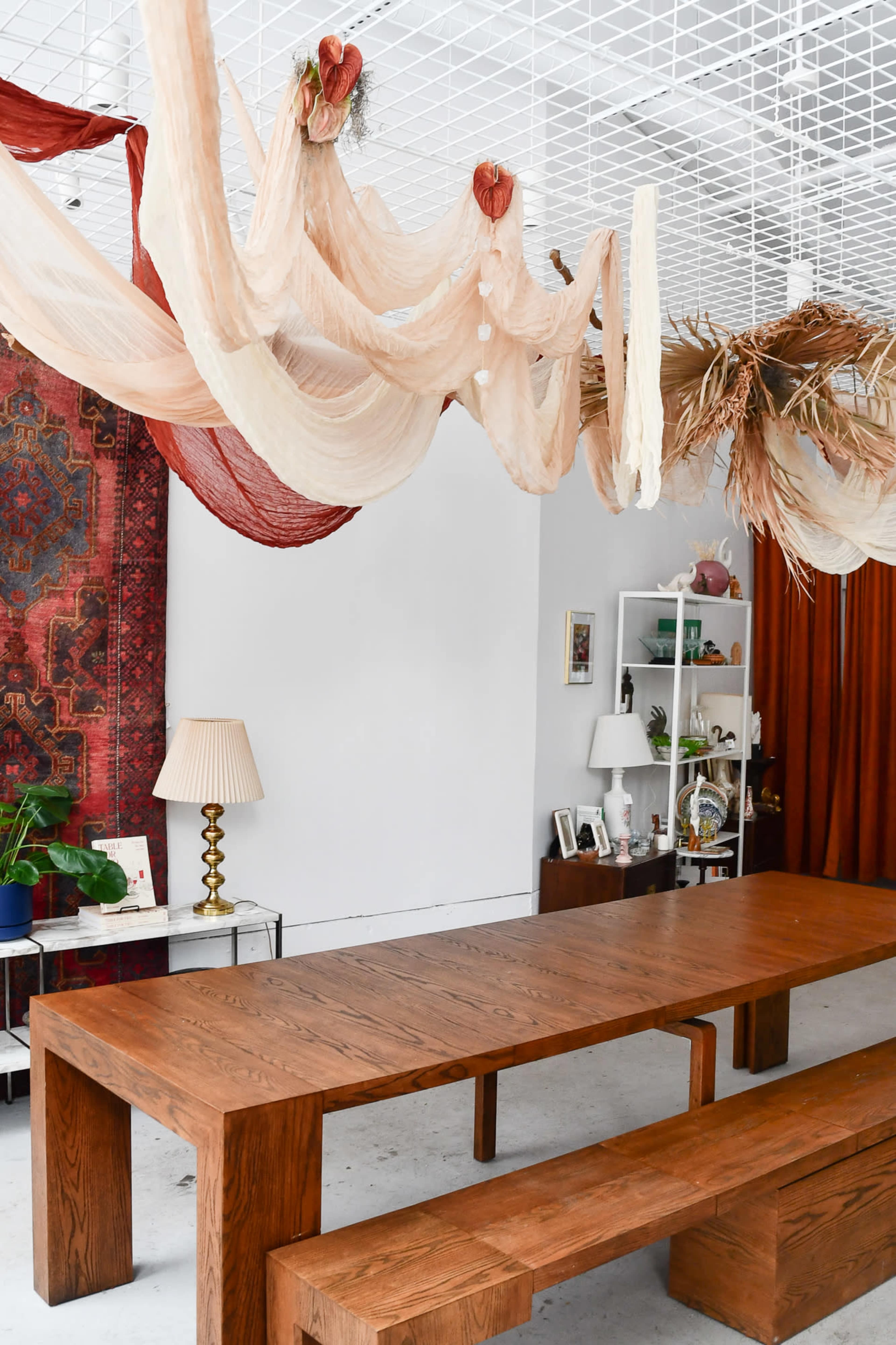 The image shows a spacious dining area with a large wooden table and bench, draped decorations hanging from the ceiling, and a patterned rug in the background.