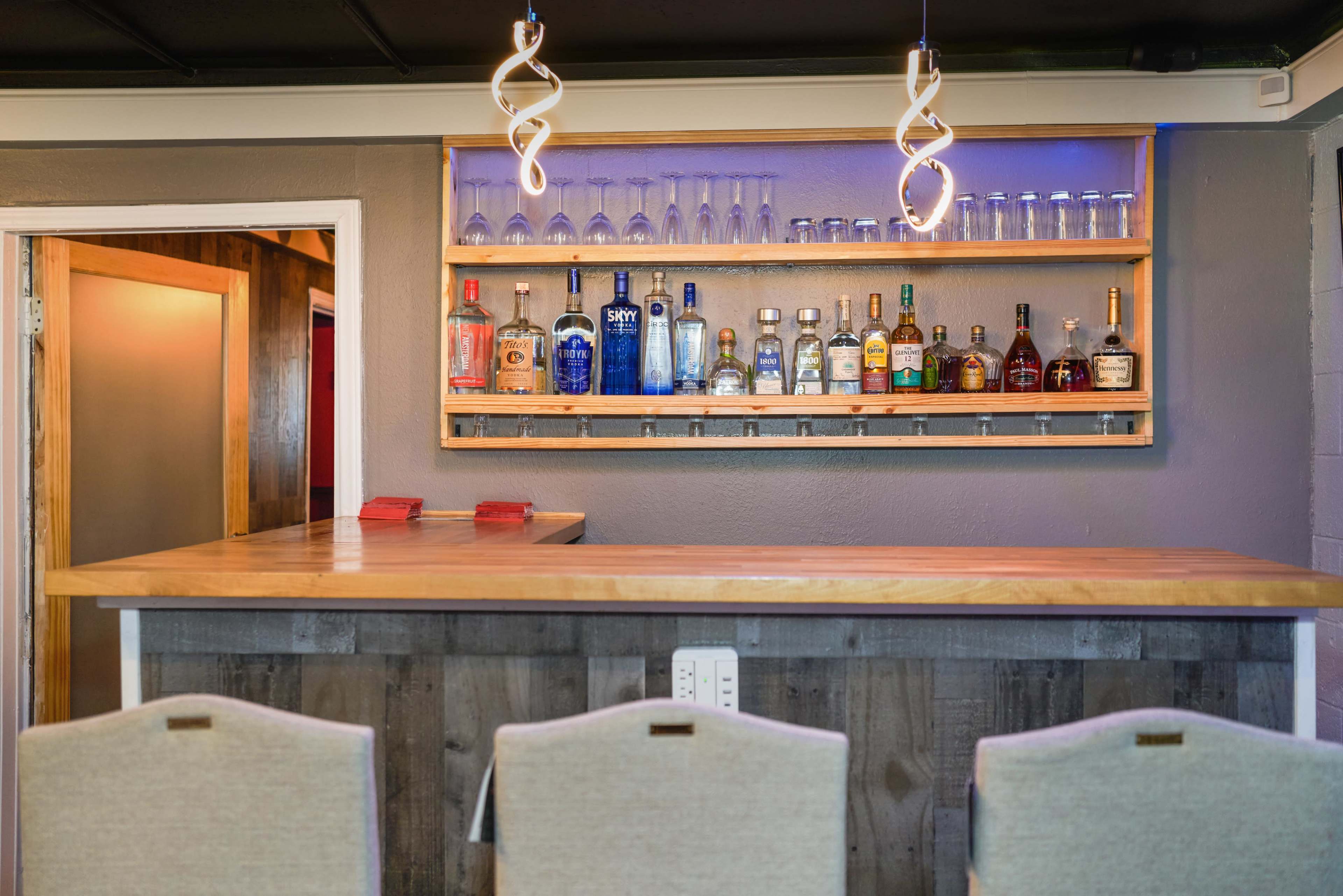A wooden bar counter with a shelf above displaying various bottles of liquor and glassware, illuminated by modern light fixtures.