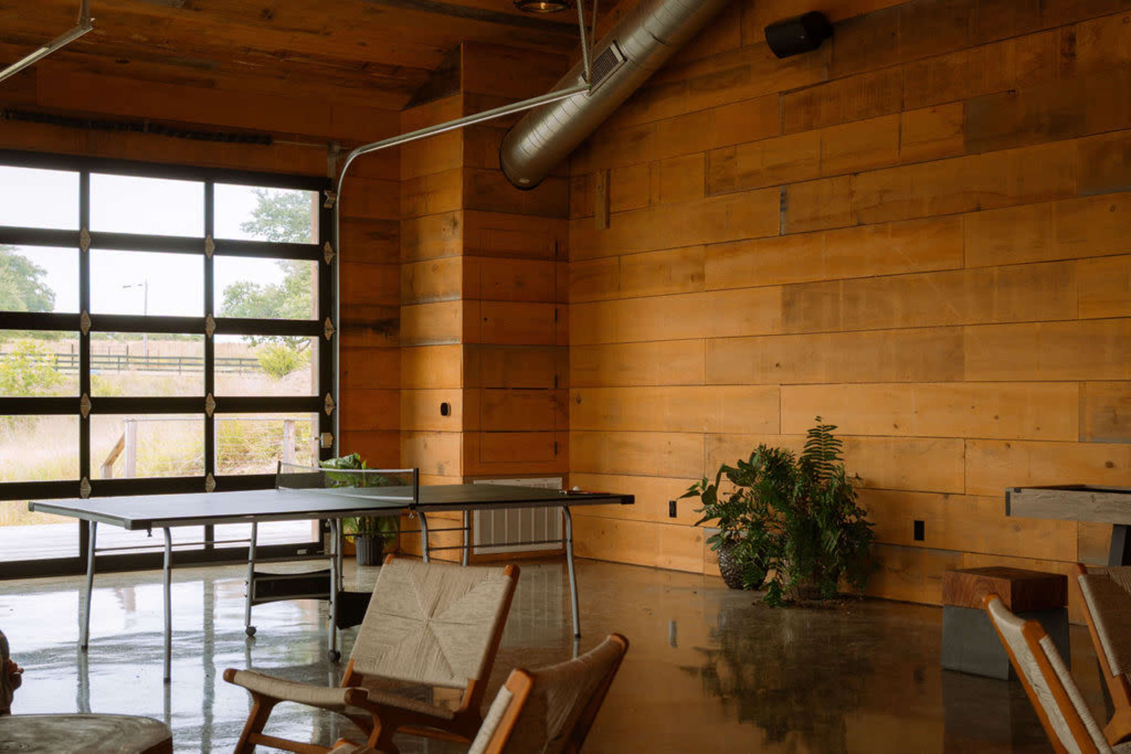 The image shows a minimalist interior space with wooden walls, a glass garage door, a potted plant, and a table set up for activities.