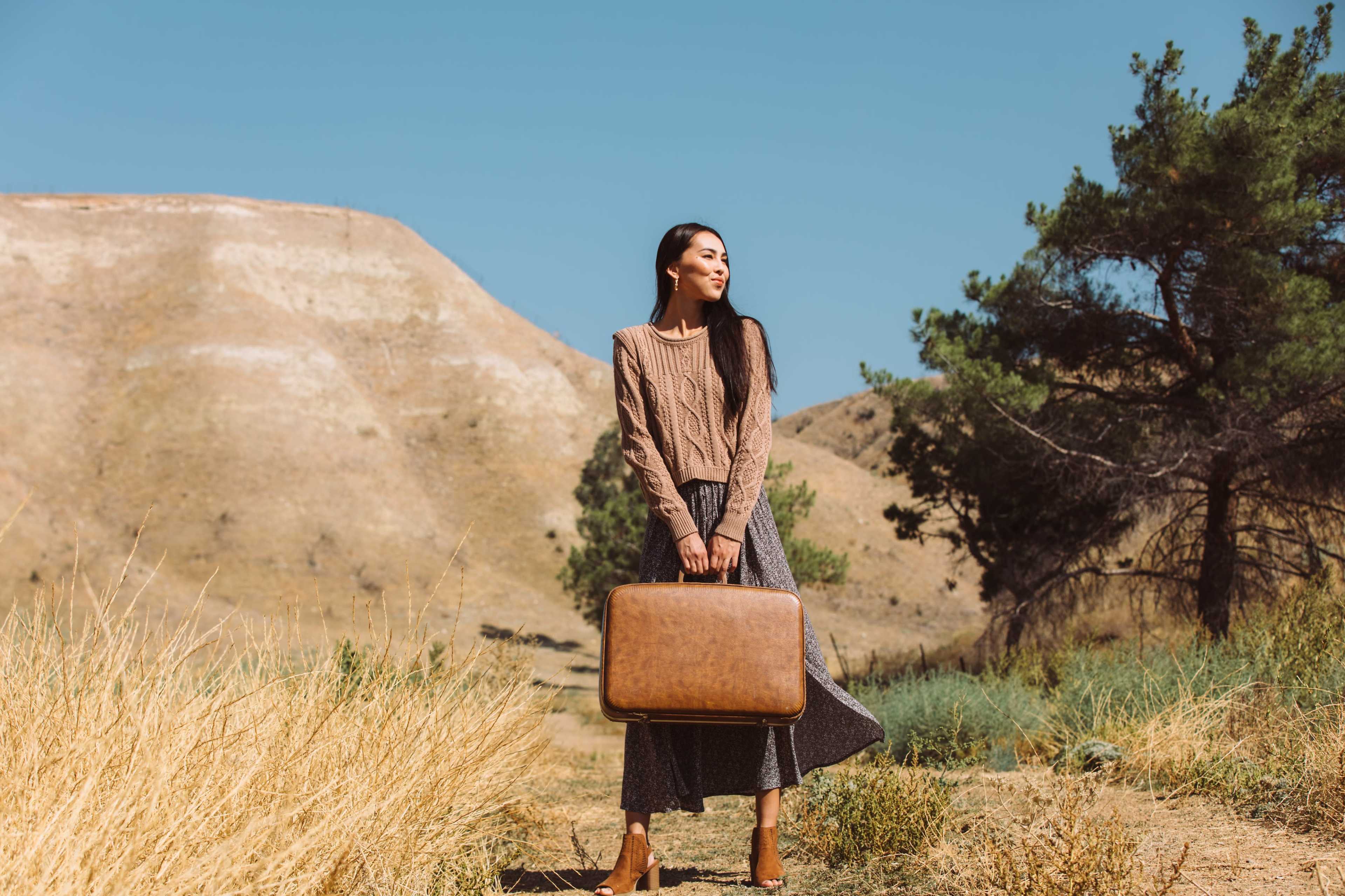 A woman stands in a dry, hilly landscape holding a brown suitcase.