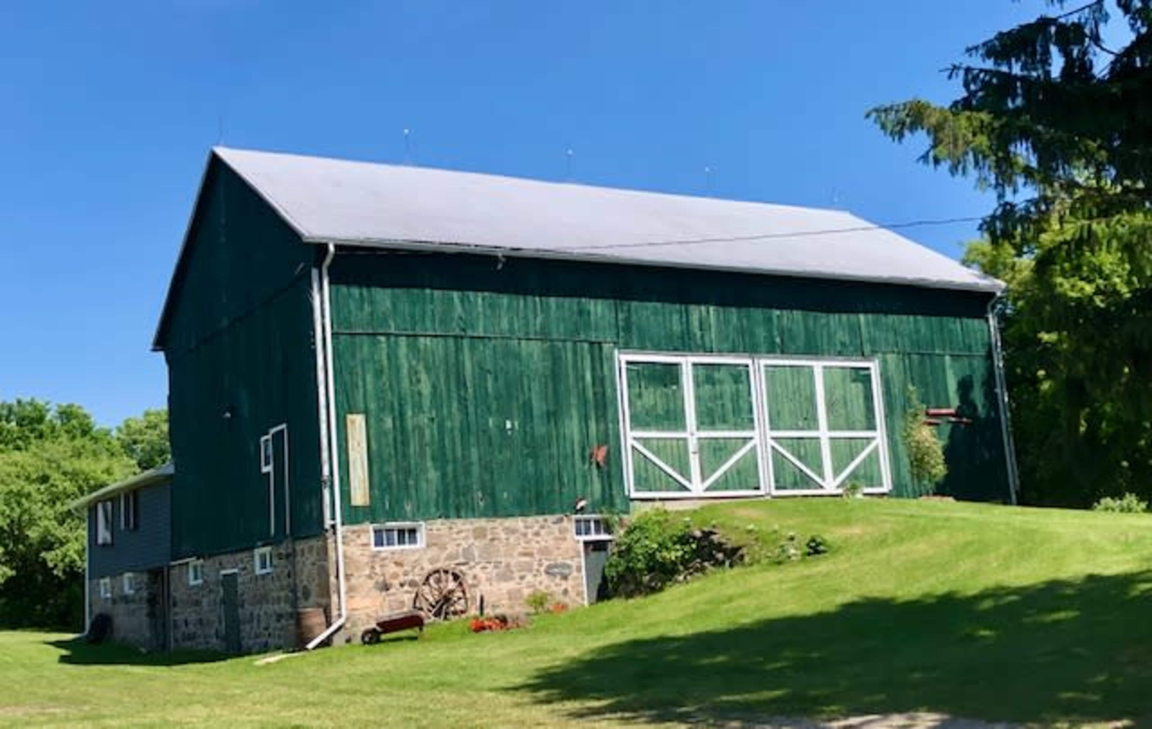 A large green barn with a stone foundation sits on a grassy slope surrounded by trees.