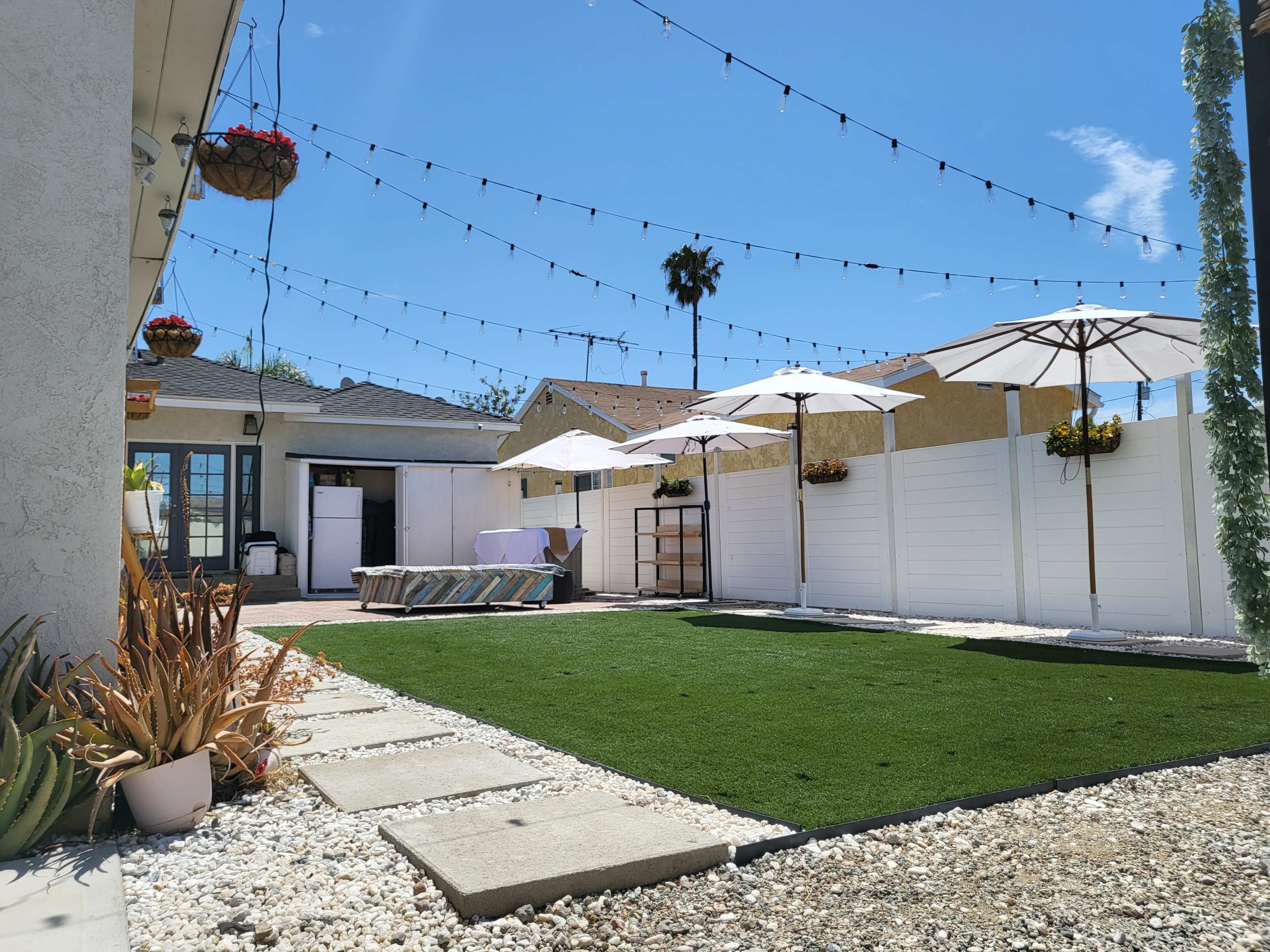 A spacious backyard with artificial green grass, white fences, hanging flower baskets, umbrellas, and string lights overhead.