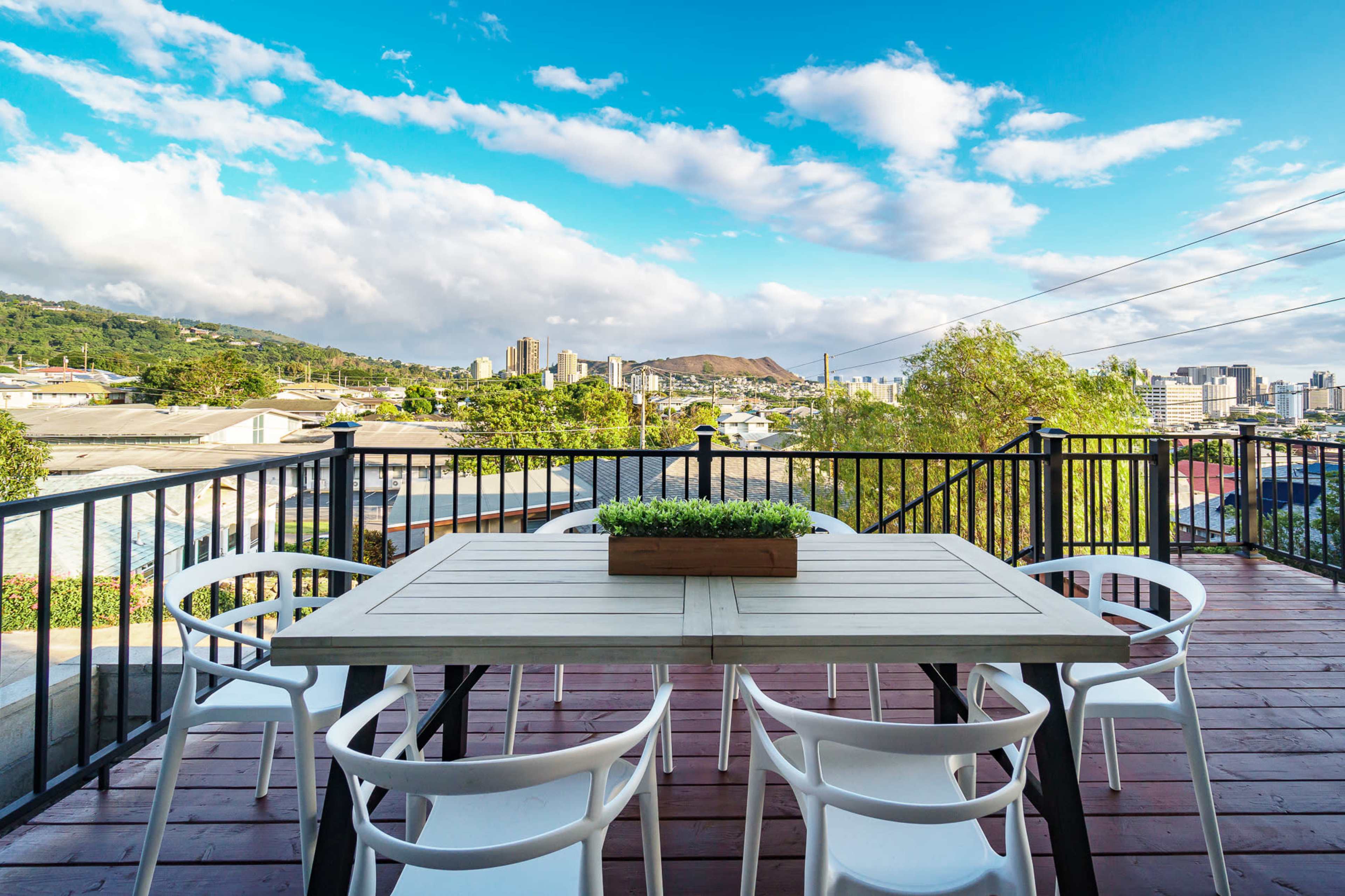 A modern outdoor dining table with white chairs overlooks a cityscape and mountains under a partly cloudy sky.