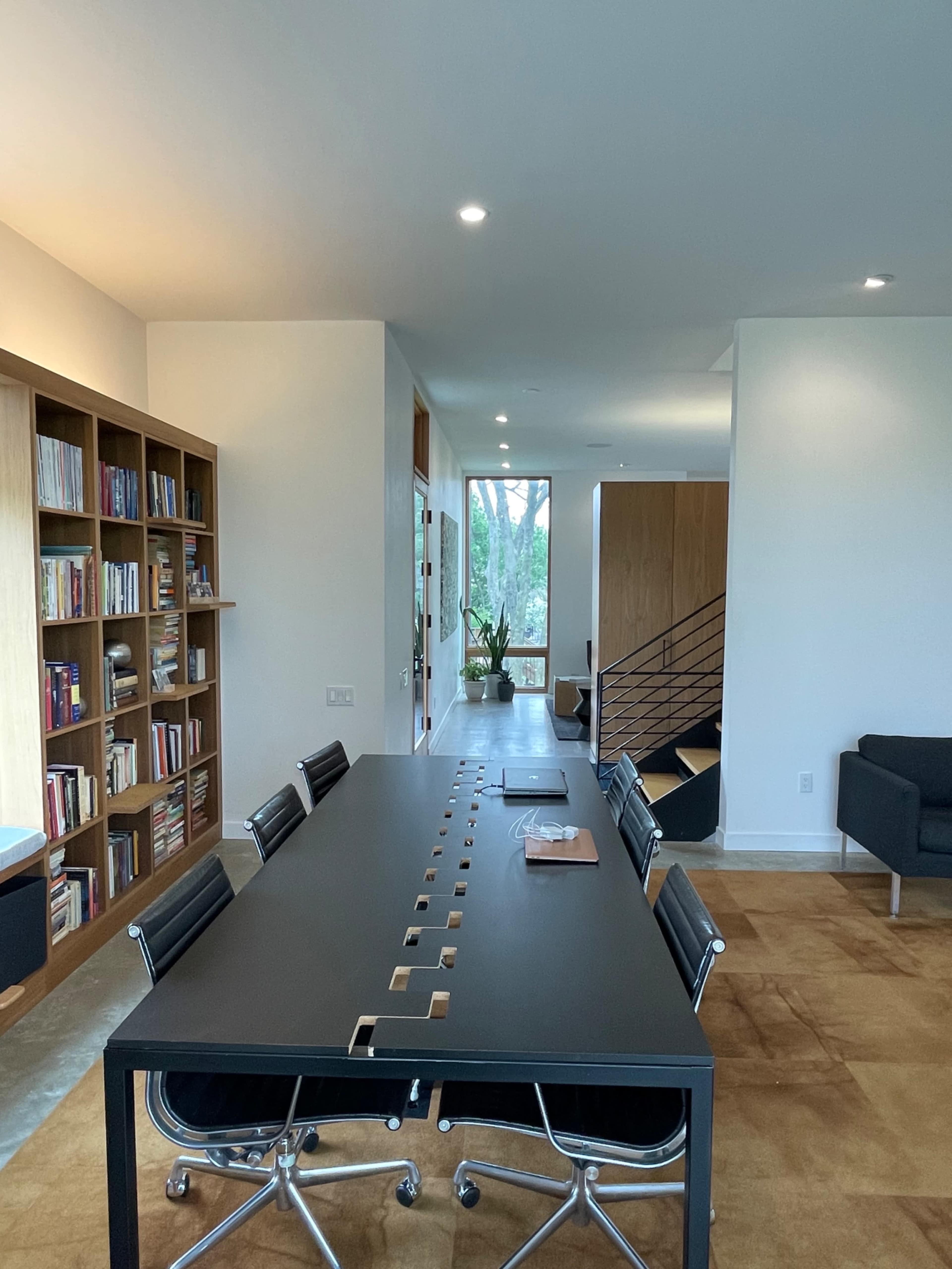 A modern office space features a long table surrounded by chairs, with a bookshelf on the left and large windows letting in natural light.