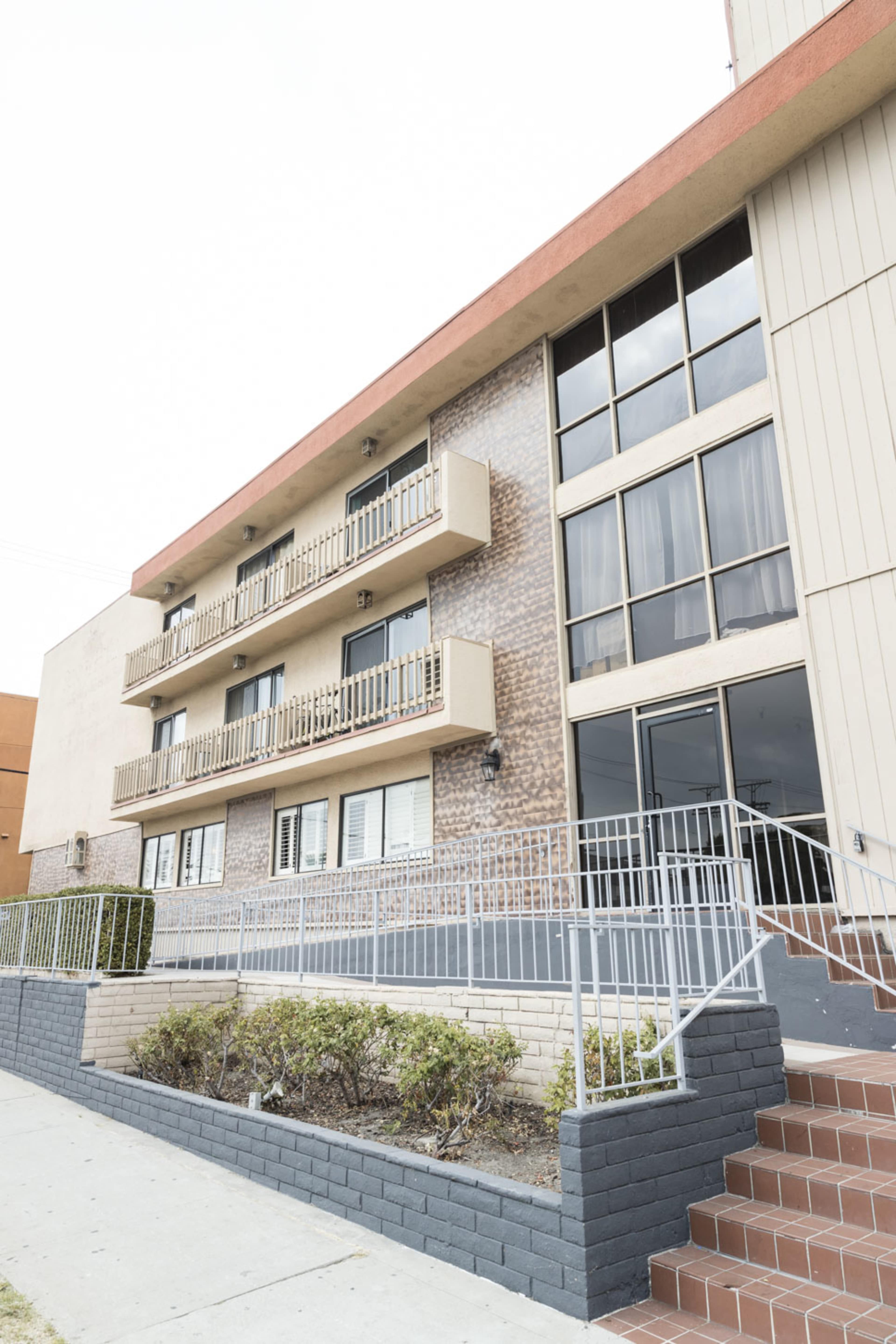 The image shows a multi-story apartment building with a brick exterior, balconies, and a glass entrance.