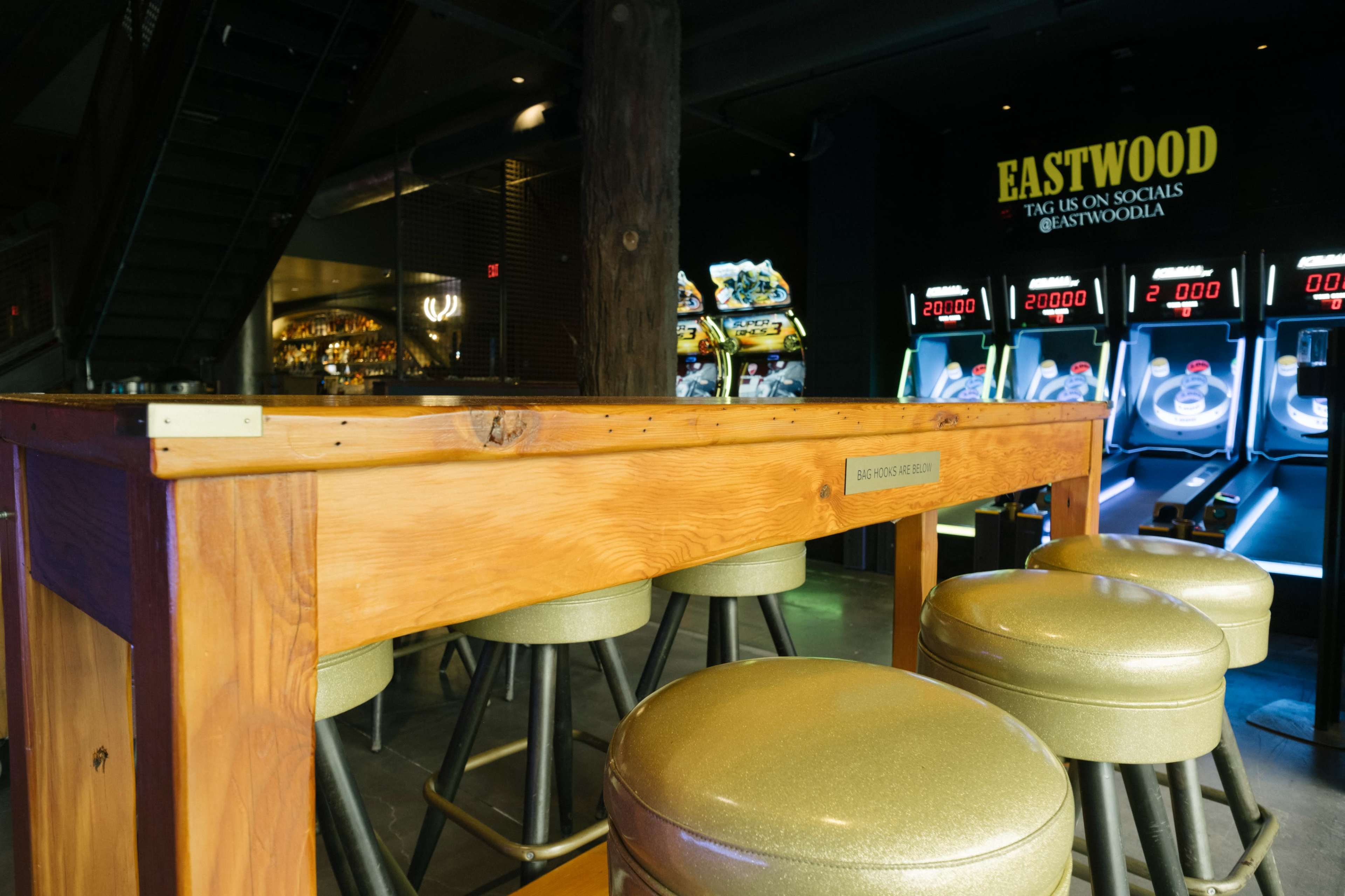 The image shows a wooden bar table with gold-colored stools in a dimly lit venue featuring gaming machines in the background.