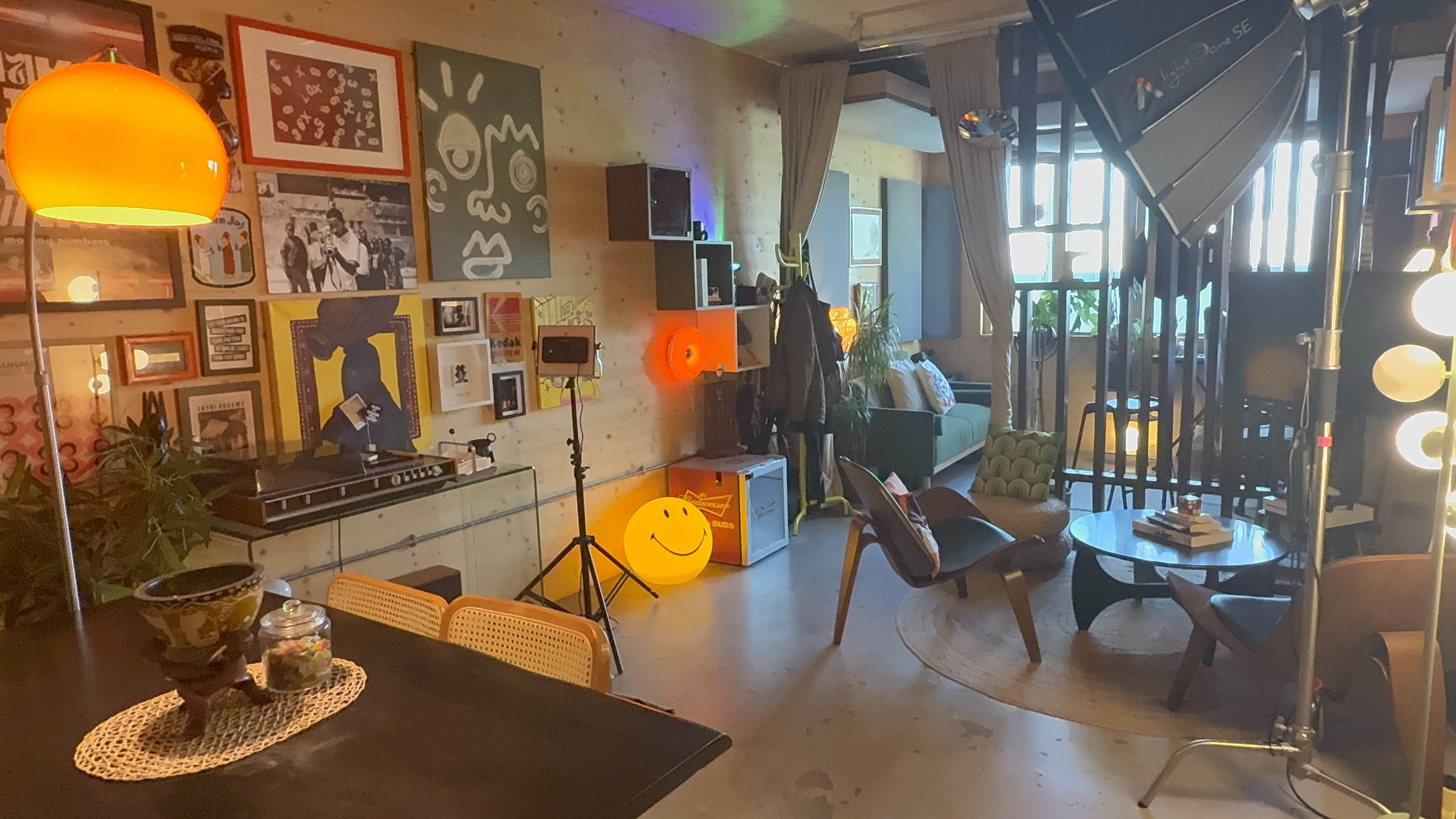 The image shows a vibrant interior space with a mix of furniture, colorful artwork on the walls, and a cozy seating area illuminated by various light sources.