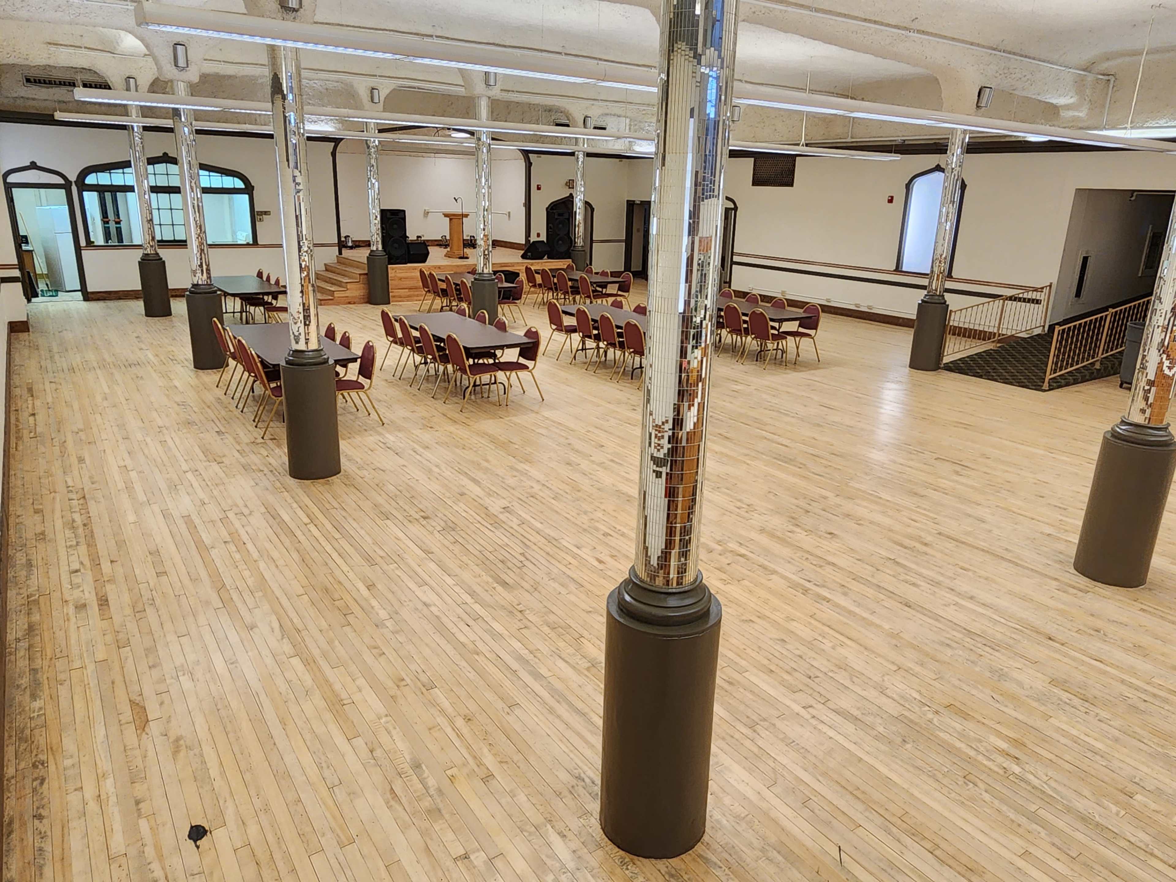 The image shows a spacious indoor venue with polished wooden floors, arranged seating around tables, and decorative columns supporting the ceiling.