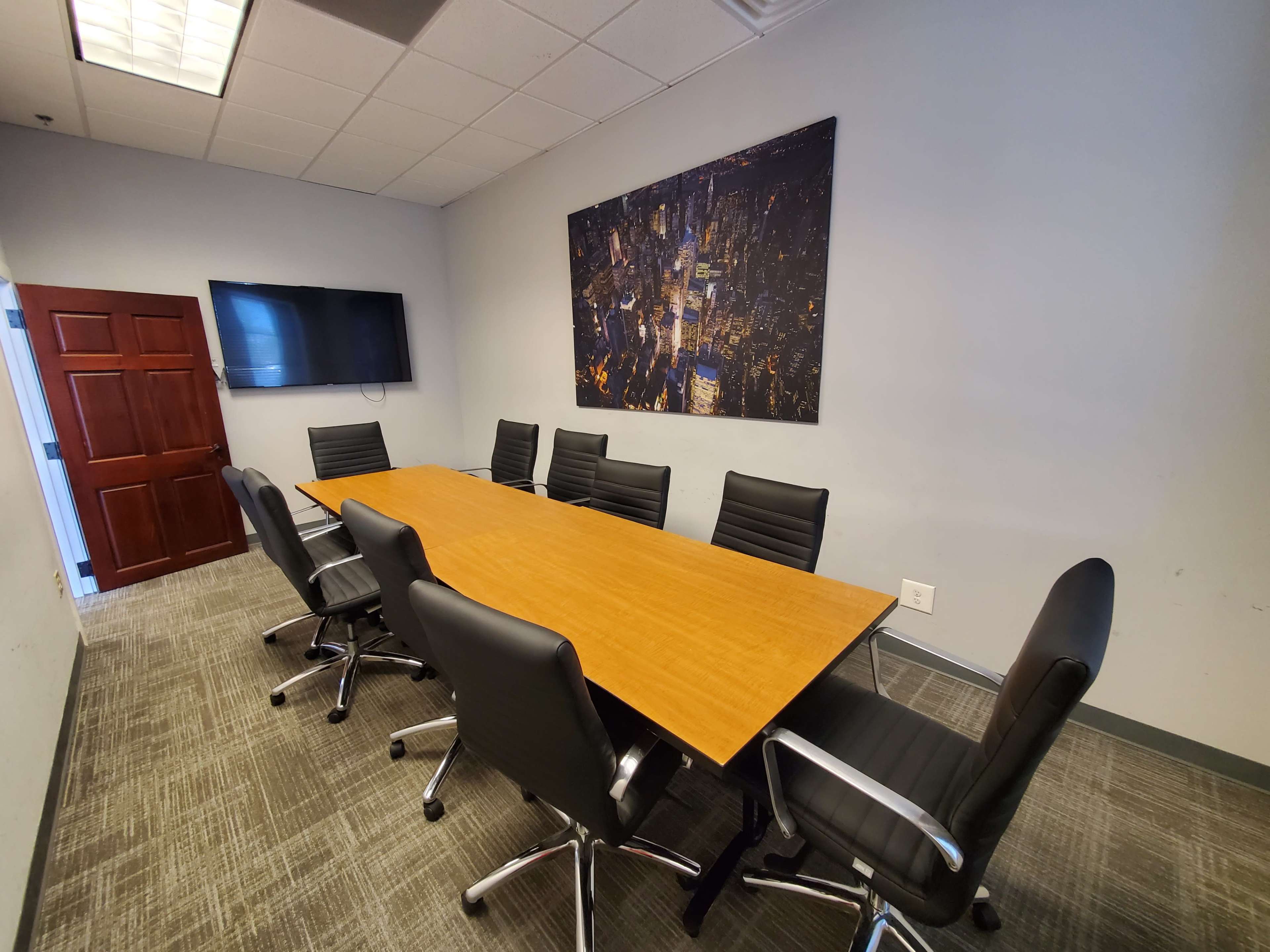 A modern conference room features a large wooden table surrounded by black leather chairs, with a television on the wall and a cityscape artwork.