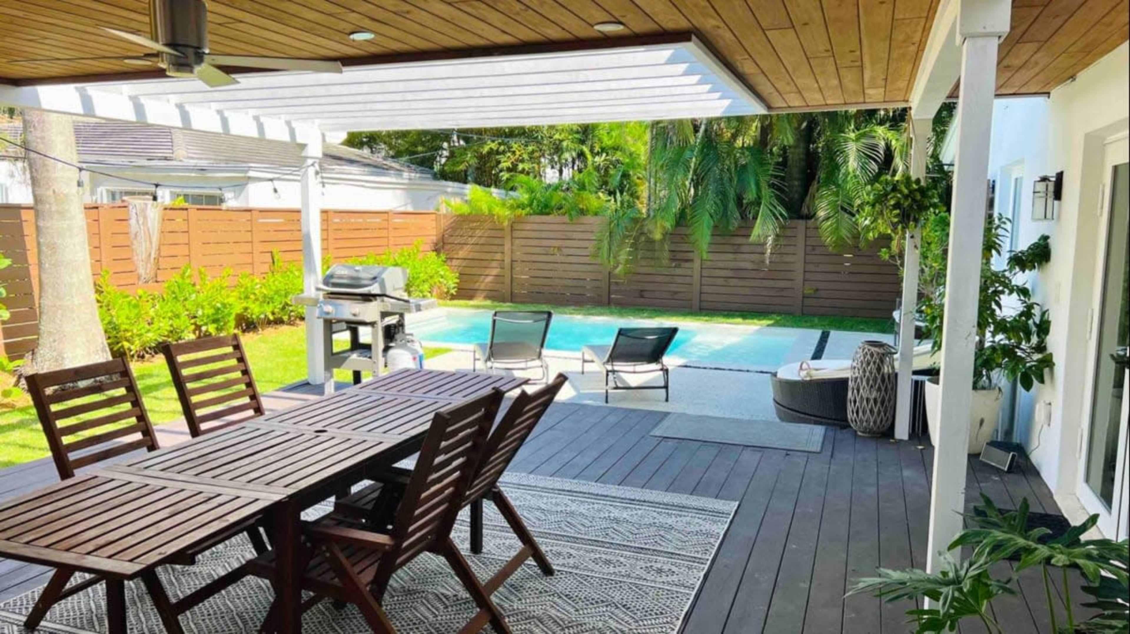 A patio area with a wooden dining table and chairs overlooking a swimming pool surrounded by greenery.