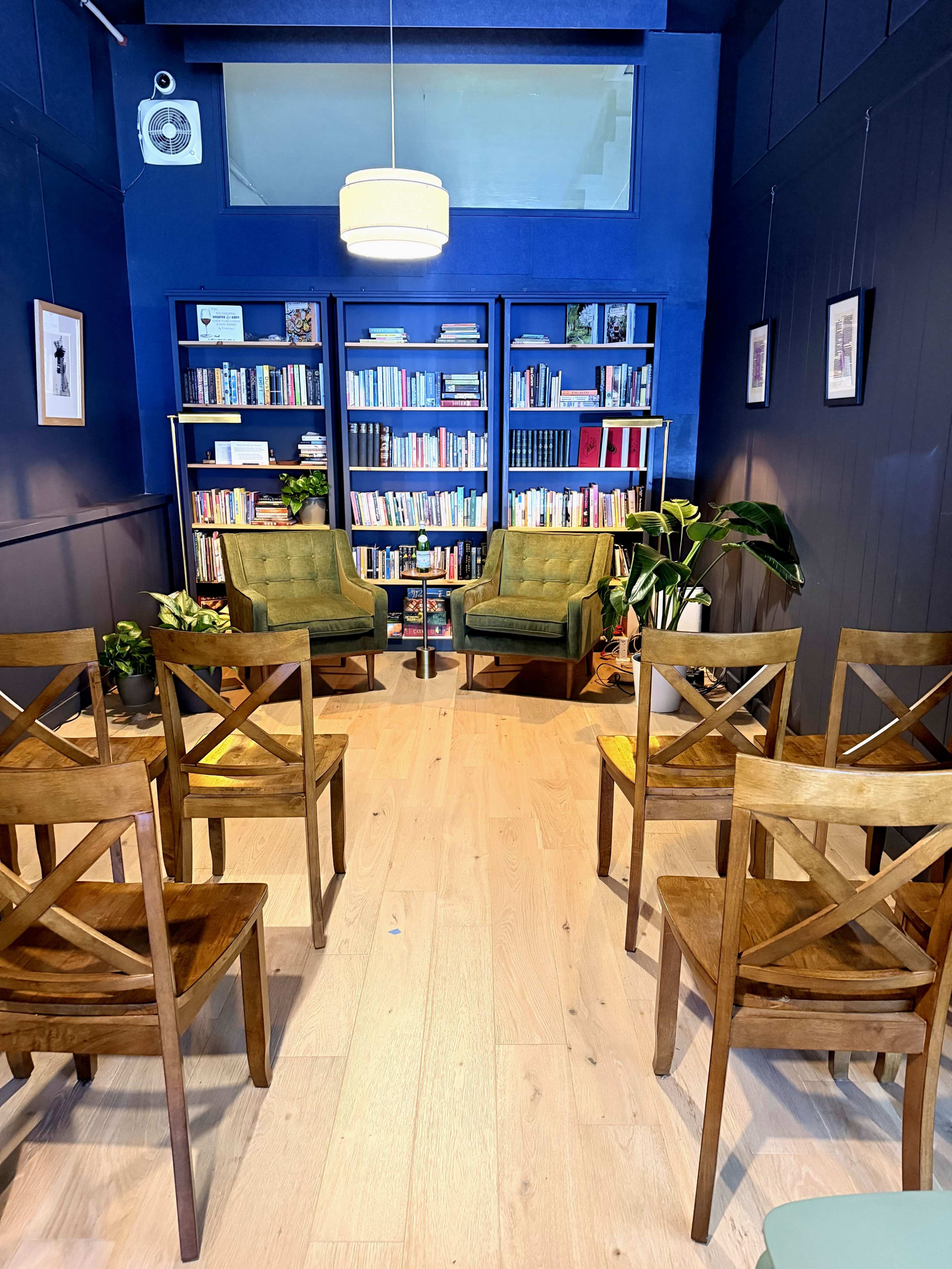 The image shows a cozy reading nook with two green armchairs facing each other, surrounded by bookshelves filled with various books, and several wooden chairs arranged in rows.