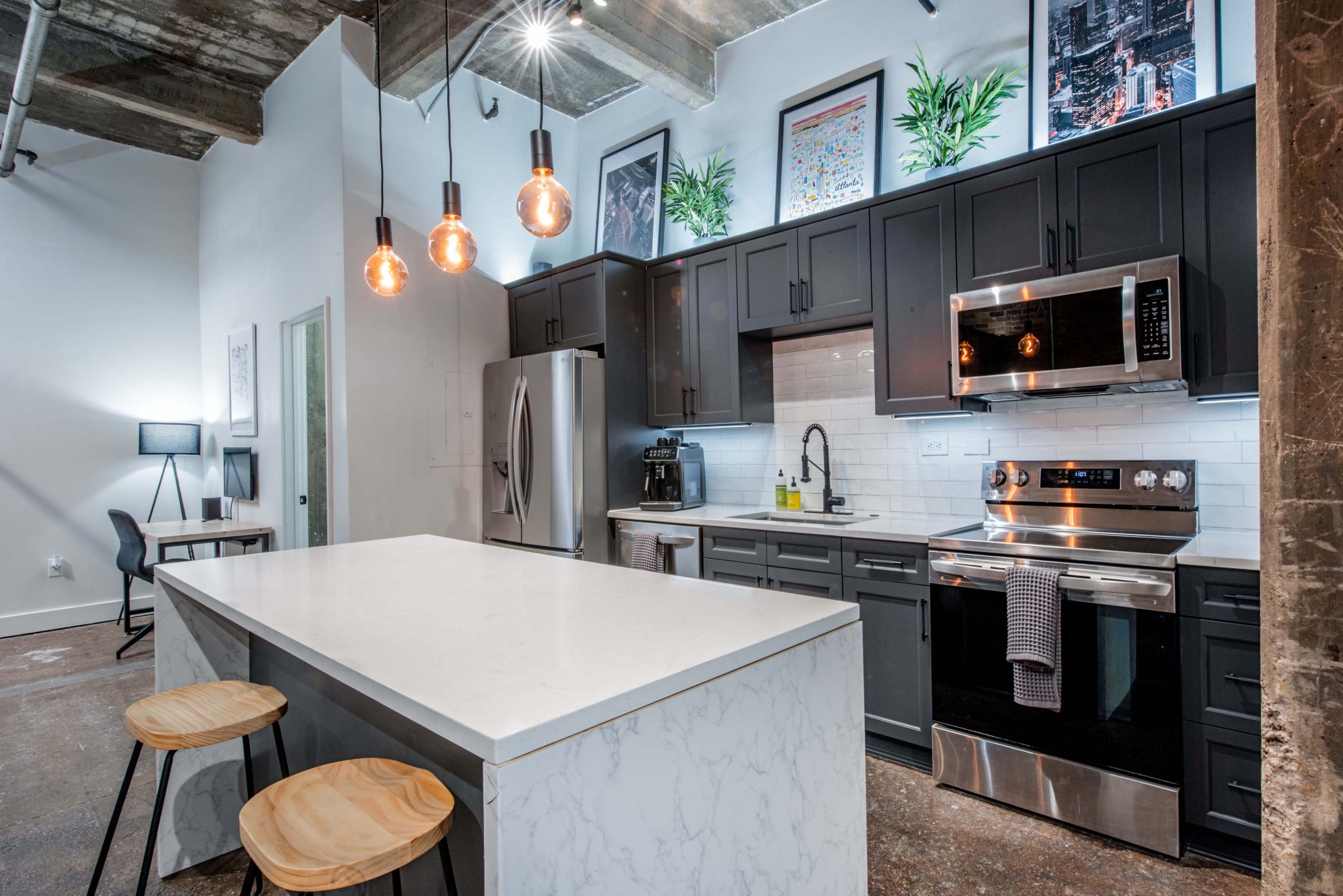 A modern kitchen featuring dark cabinetry, stainless steel appliances, a white marble island with two wooden stools, and pendant lighting.