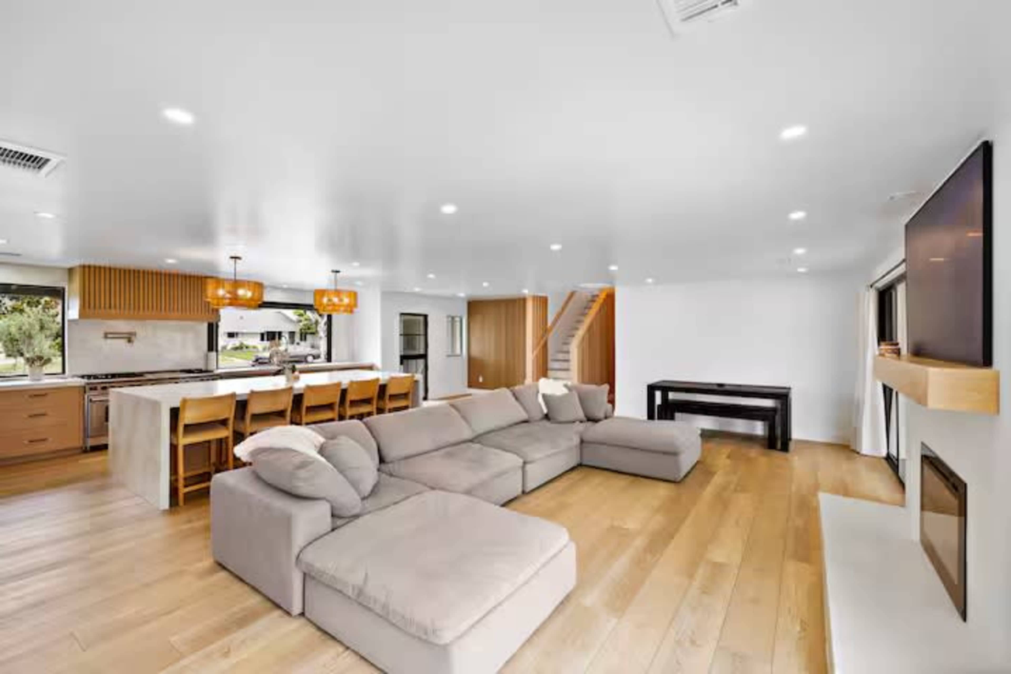 The image shows a modern living space with a large sectional sofa, an open kitchen in the background, and a staircase leading to an upper level.