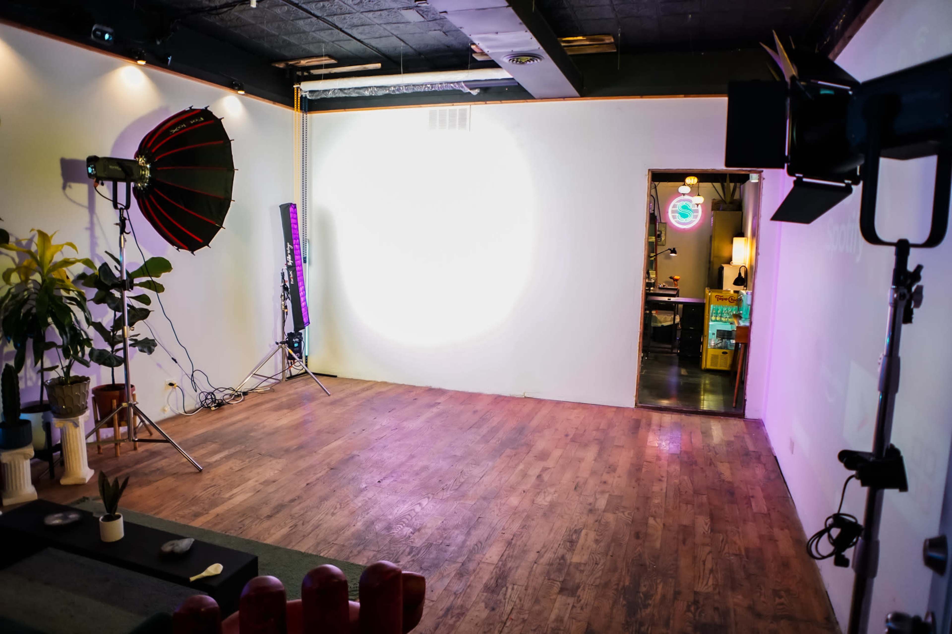 The image shows an empty studio space with a wooden floor, lighting equipment set up, and a partially visible doorway leading to another area.
