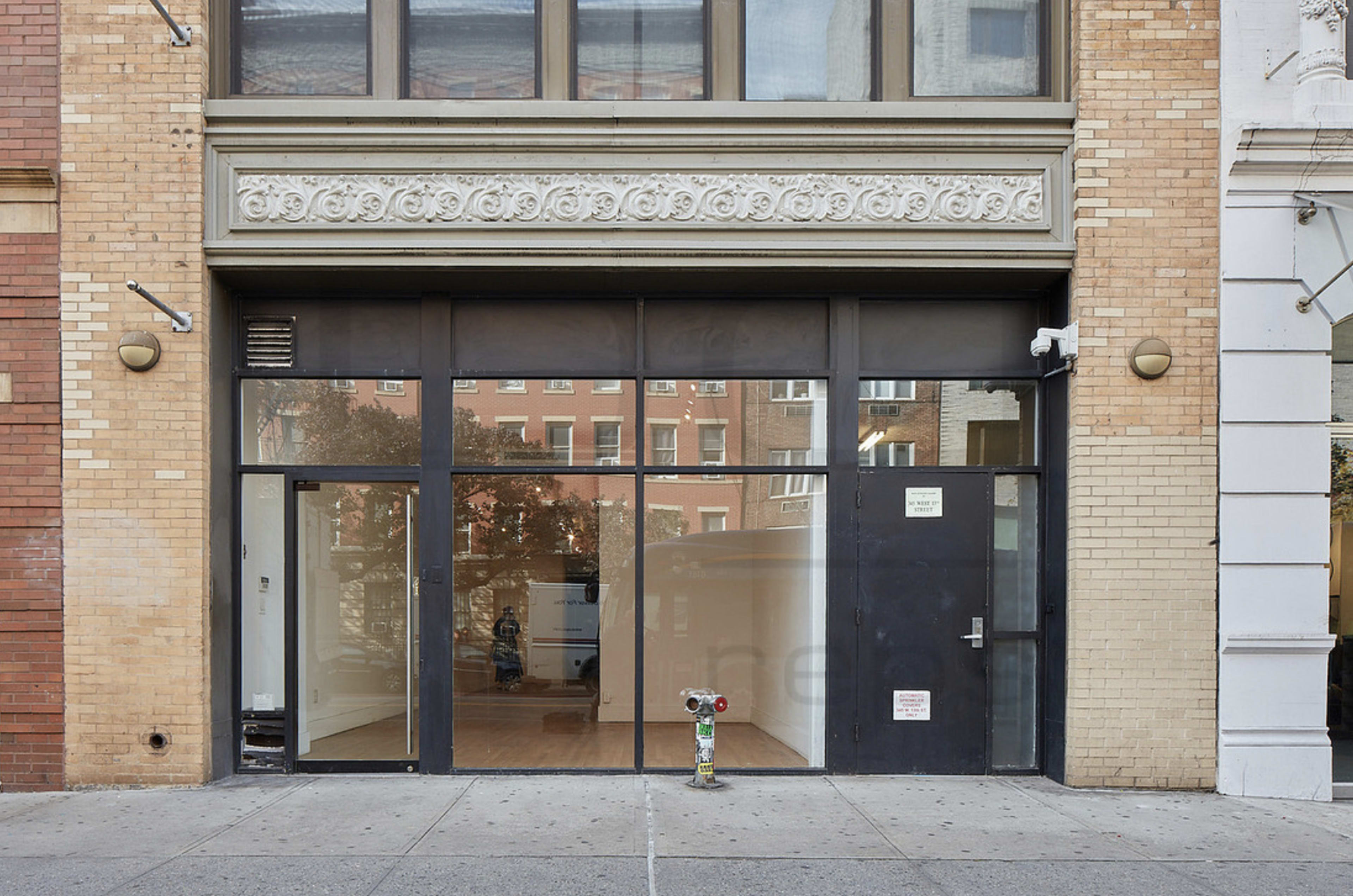 The image shows a large, empty storefront with glass windows and a dark entrance door, flanked by brick buildings.