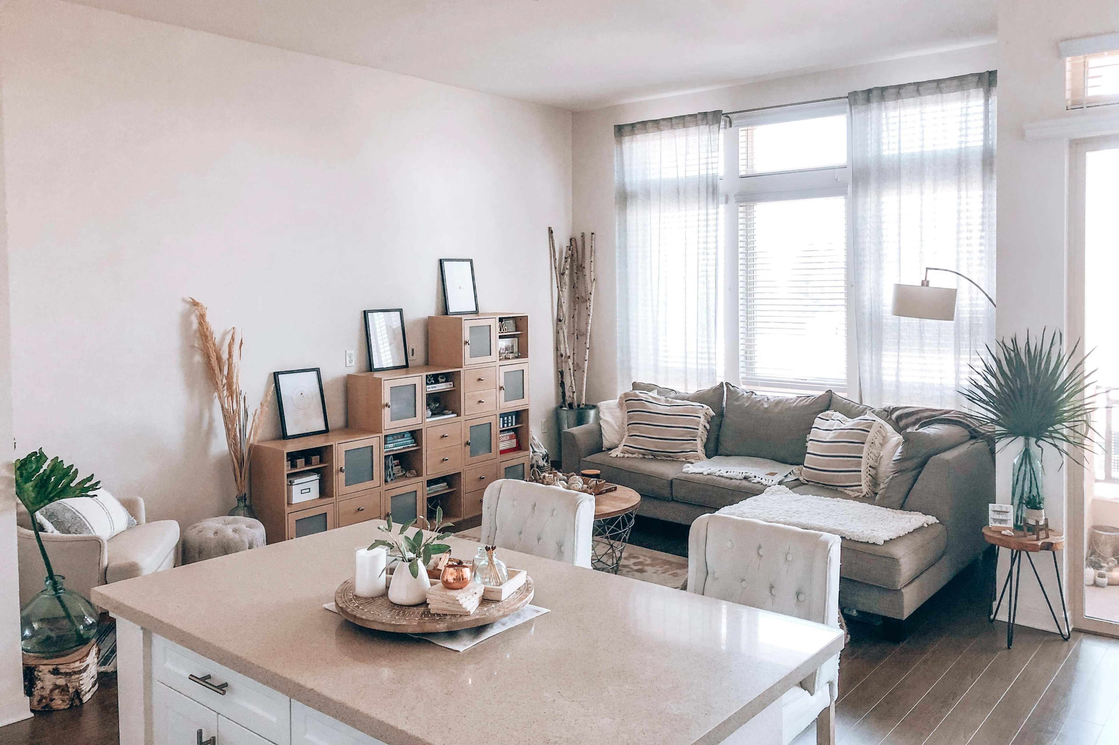 A modern living room features a sectional sofa, a bookshelf, and a dining table, with large windows allowing natural light to enter.