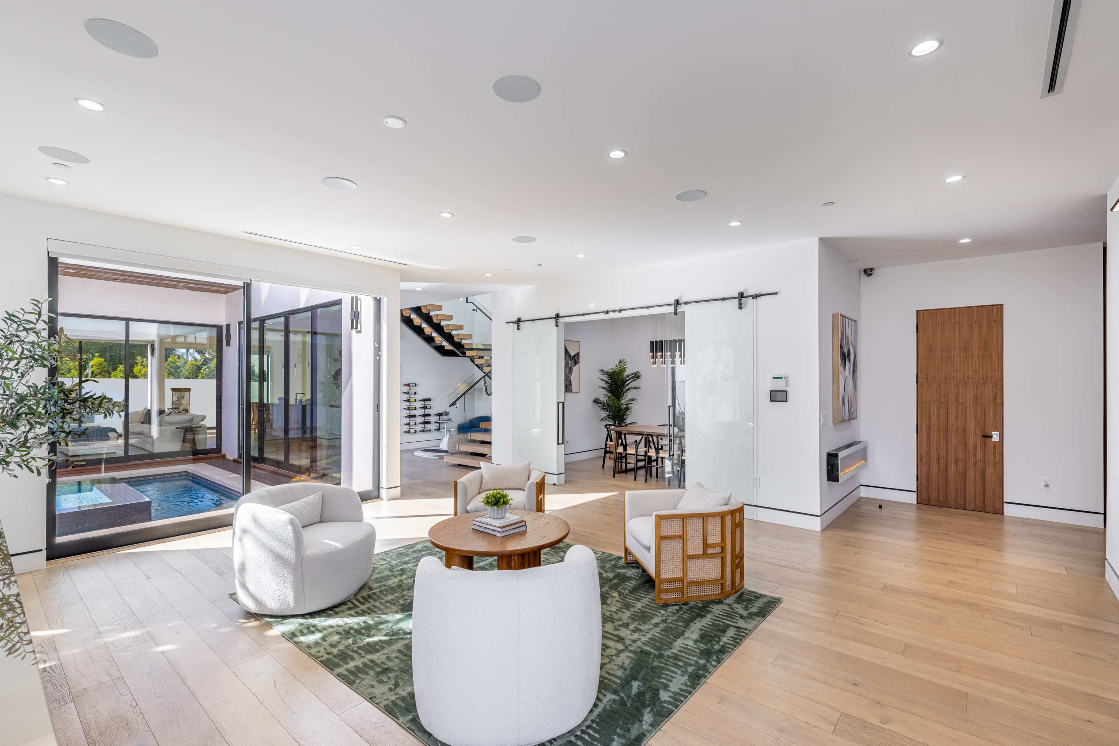 A modern living room with white furniture, large windows leading to an outdoor space, and a staircase in the background.