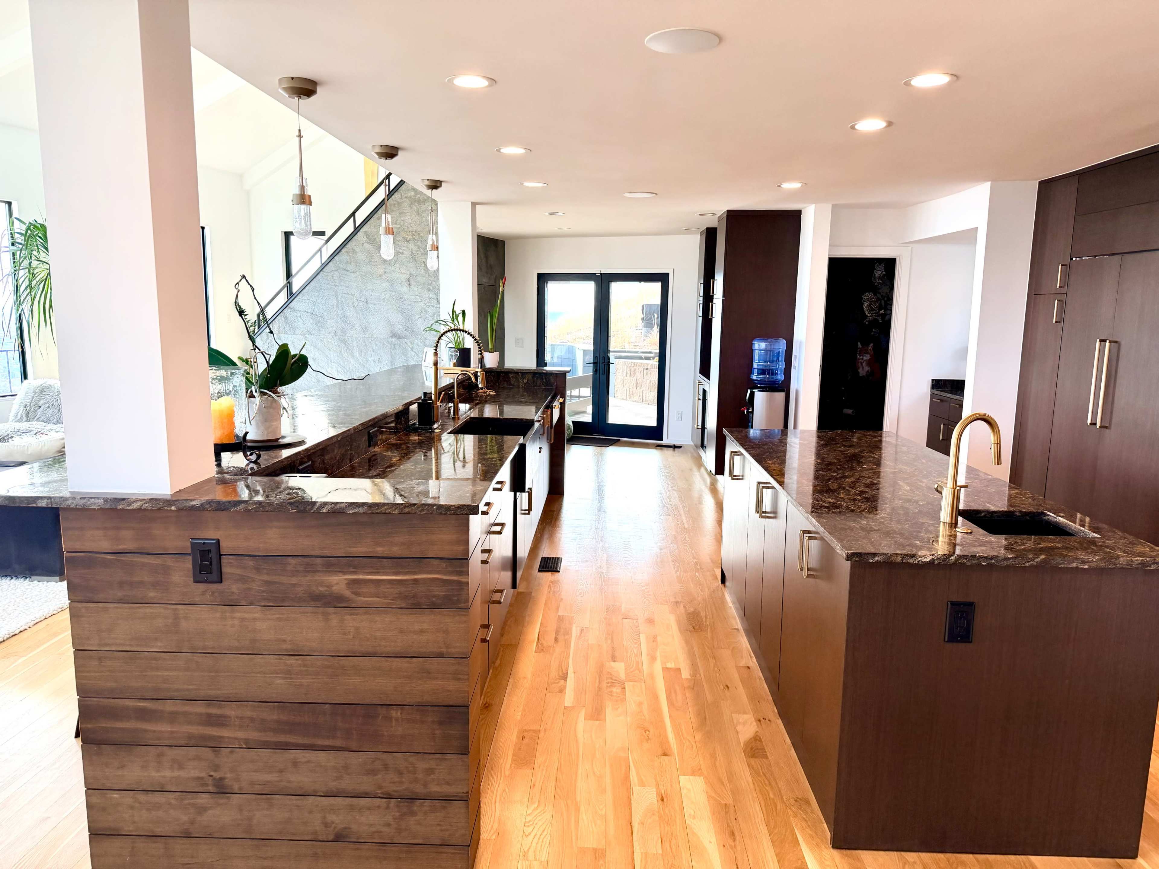 The image shows a modern kitchen with two large islands featuring dark countertops and wooden cabinetry, connected to a spacious living area.
