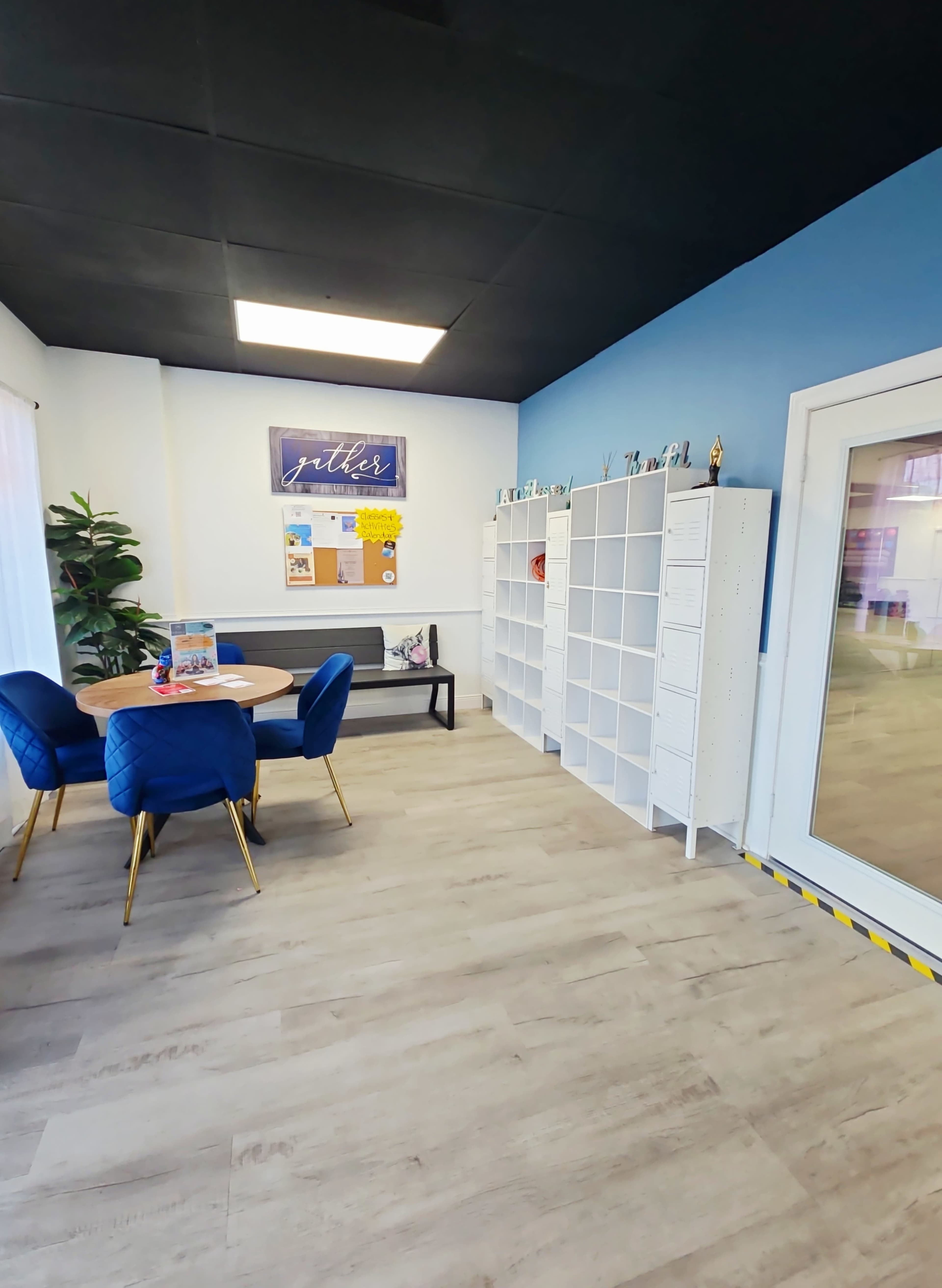 The image shows a modern office space with blue walls, a round wooden table surrounded by blue chairs, and a white shelving unit along one wall.