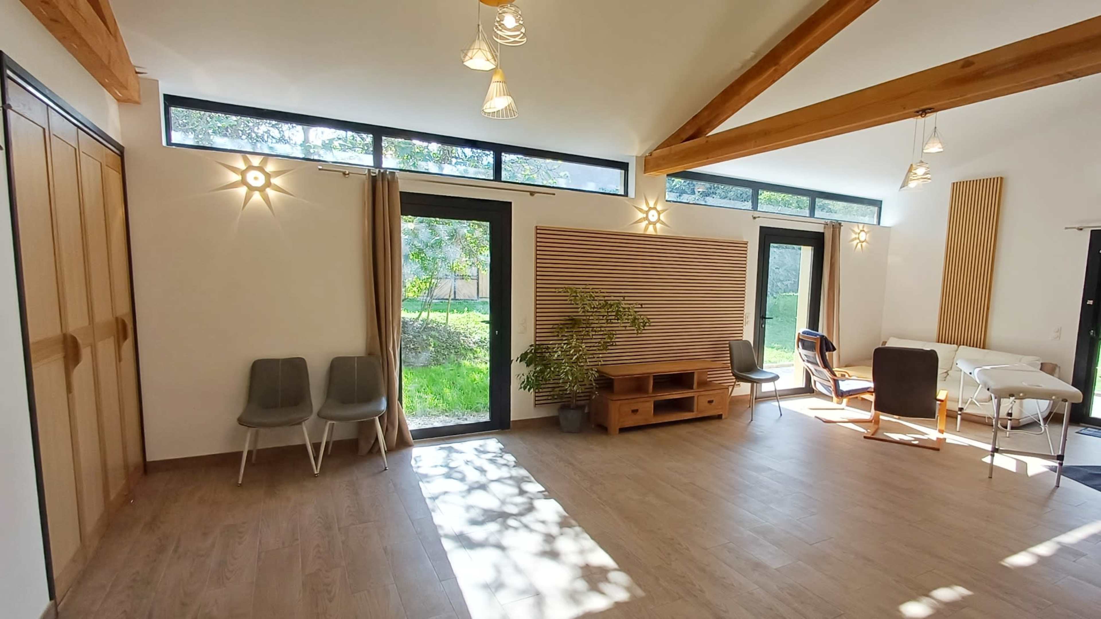 The image shows a spacious, well-lit room with large windows, wooden beams, and simple furniture including chairs and a table.