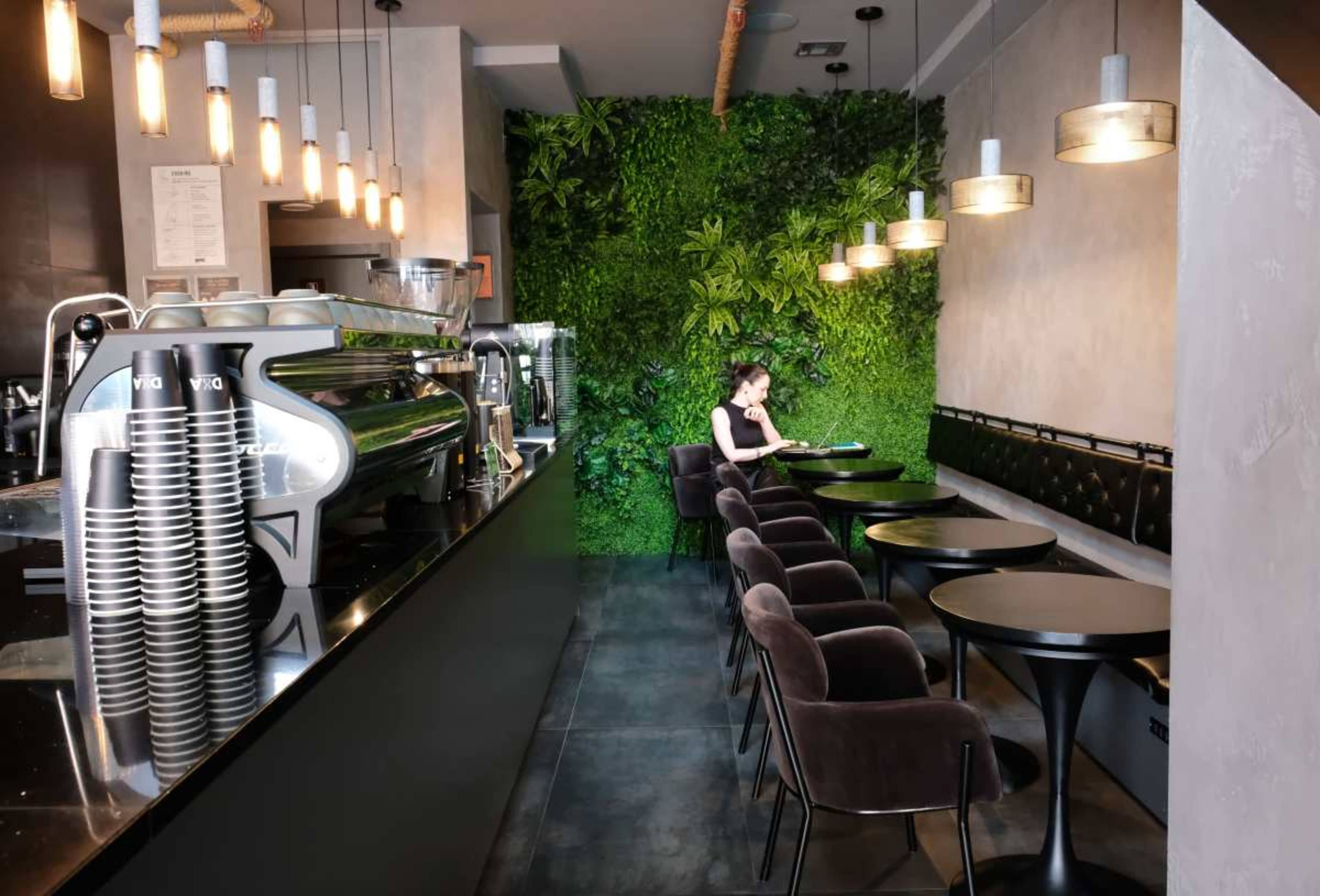 A woman is seated at a table in a modern café with a greenery wall and dark furnishings, while an espresso machine is visible on the counter.