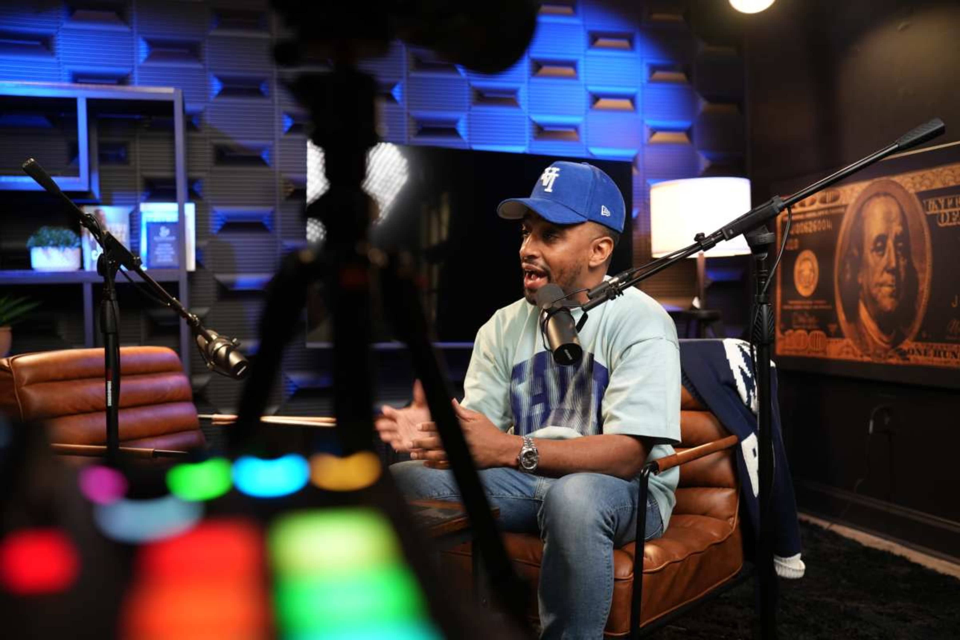 A man wearing a blue cap and light blue shirt is seated on a leather chair in a modern studio with microphones positioned nearby.