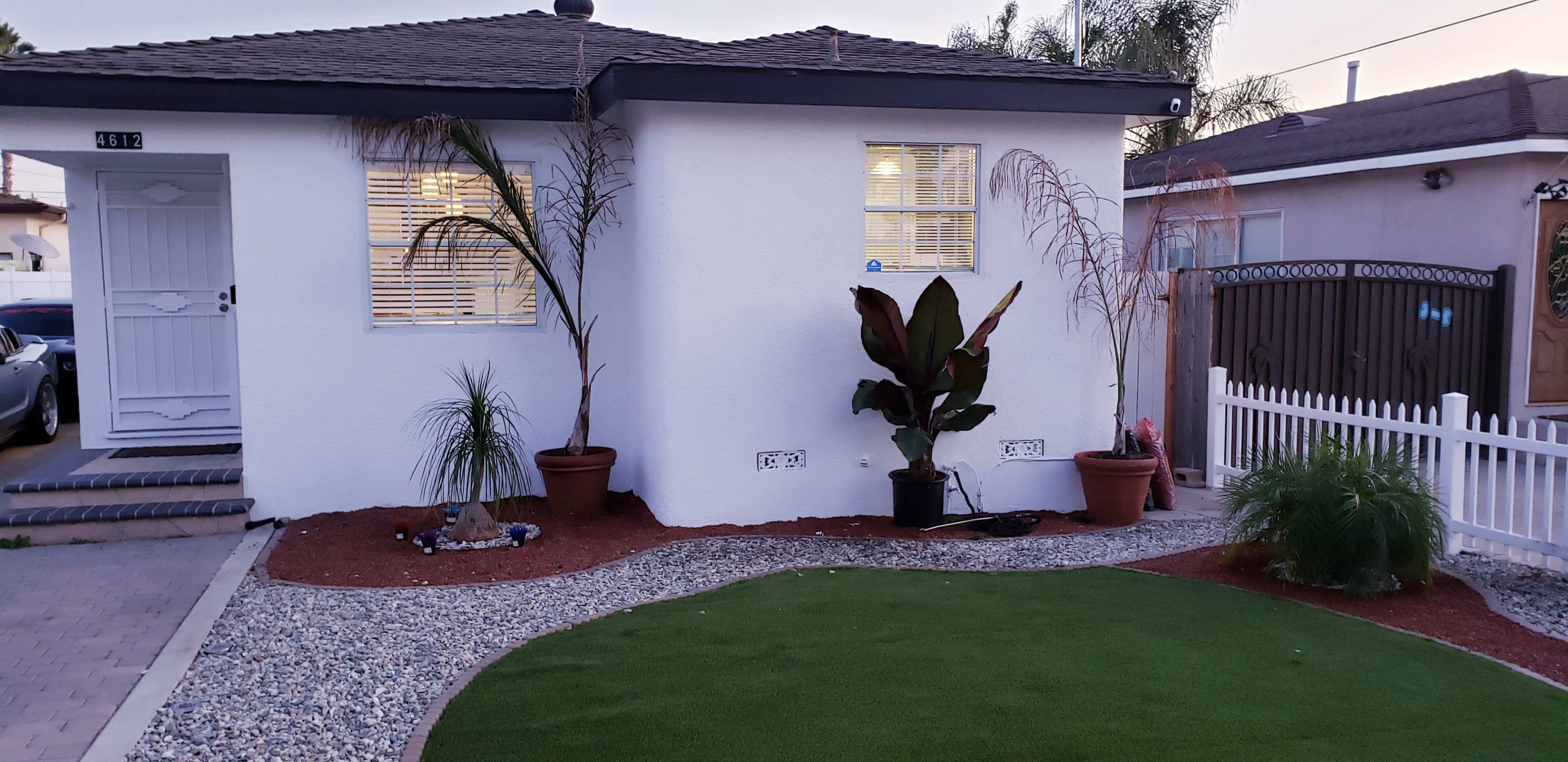 A small front yard with a white house, landscaped area featuring potted plants, gravel, and artificial grass.