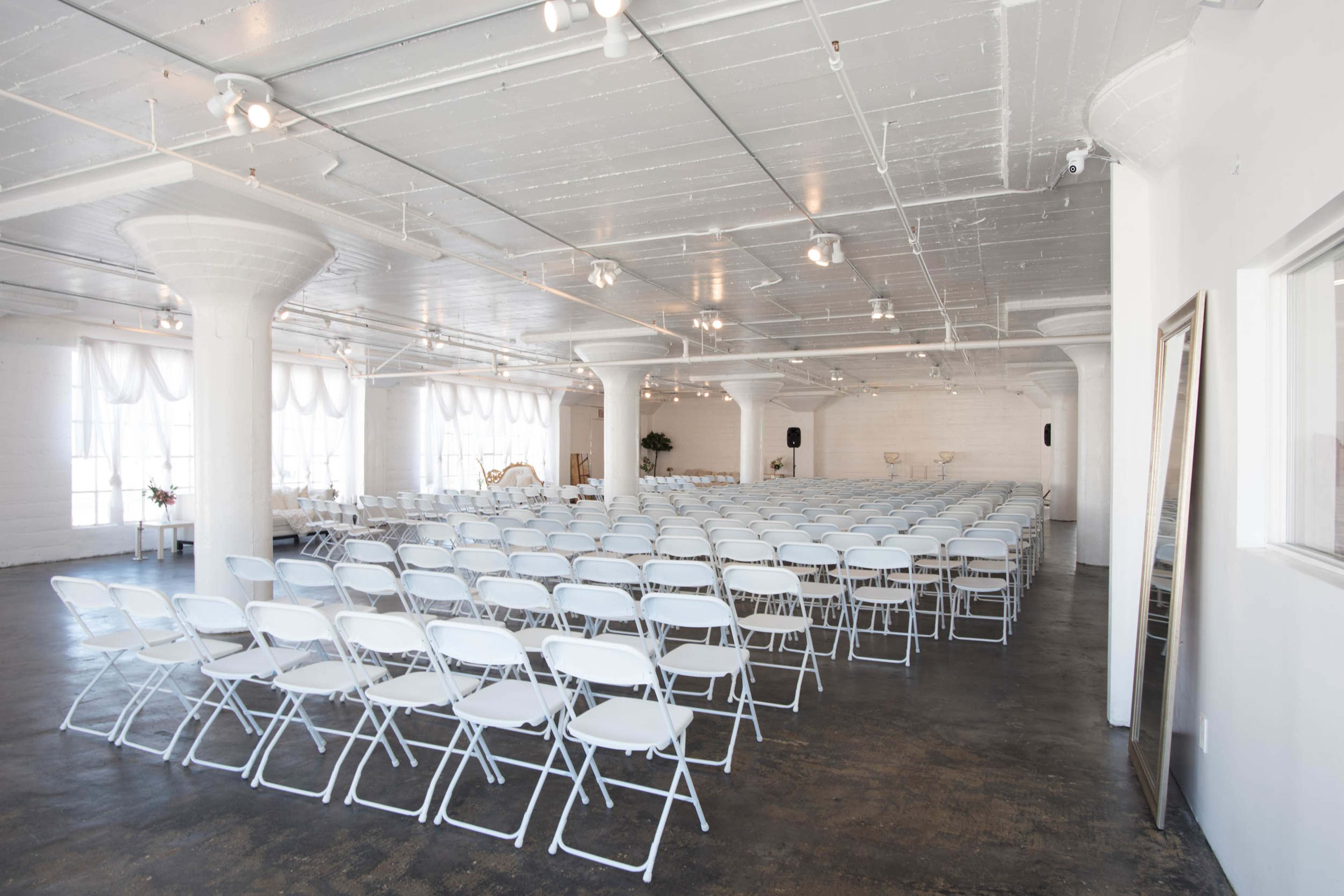 A spacious interior with rows of white folding chairs set up for an event in a brightly lit room with industrial-style columns.