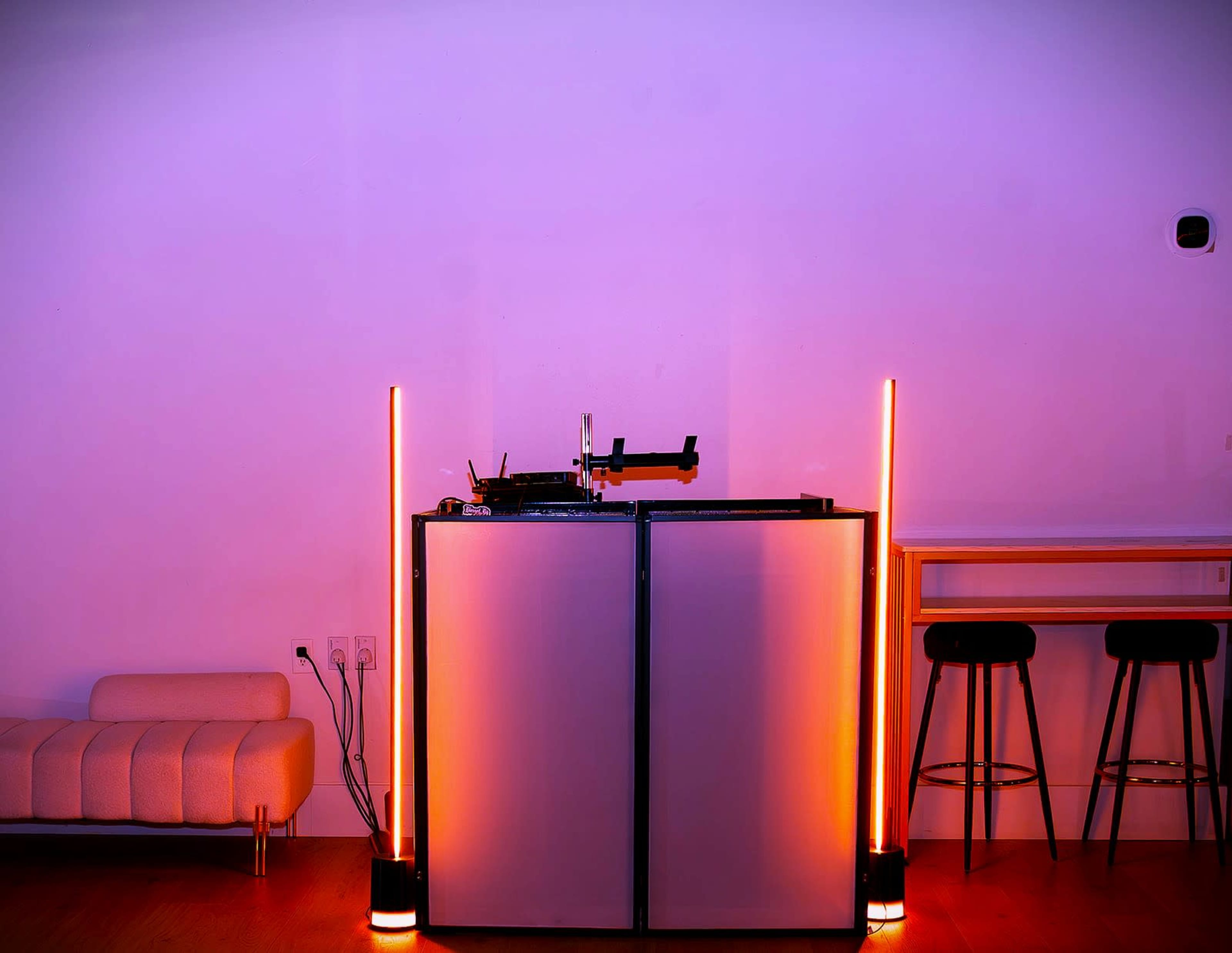 A modern entertainment setup features a turntable on a gray cabinet, flanked by glowing vertical lamps, with minimal seating and a clean white wall in the background.