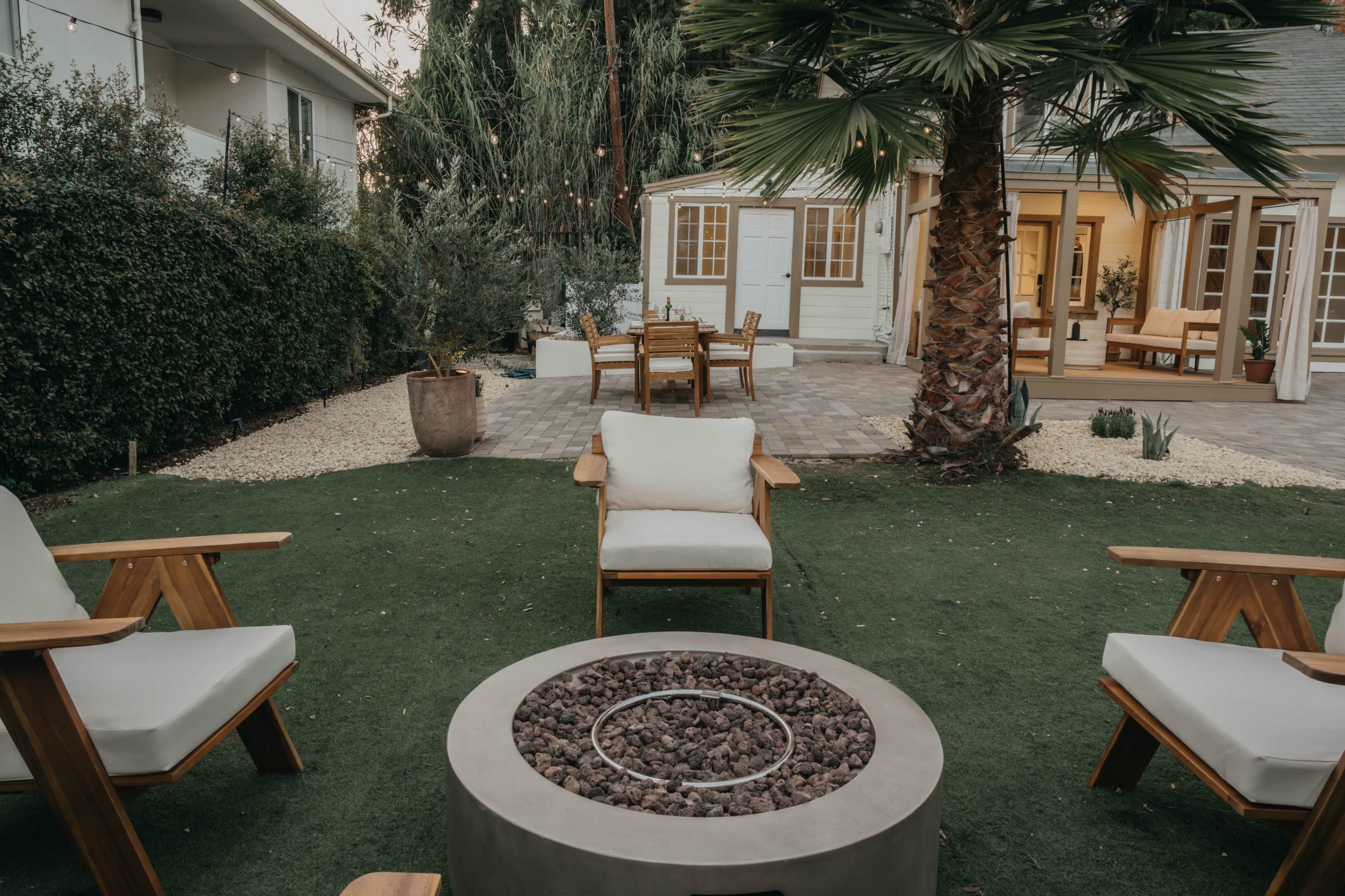 A circular fire pit surrounded by wooden chairs sits on artificial grass in a landscaped backyard with palm trees and a dining area visible in the background.