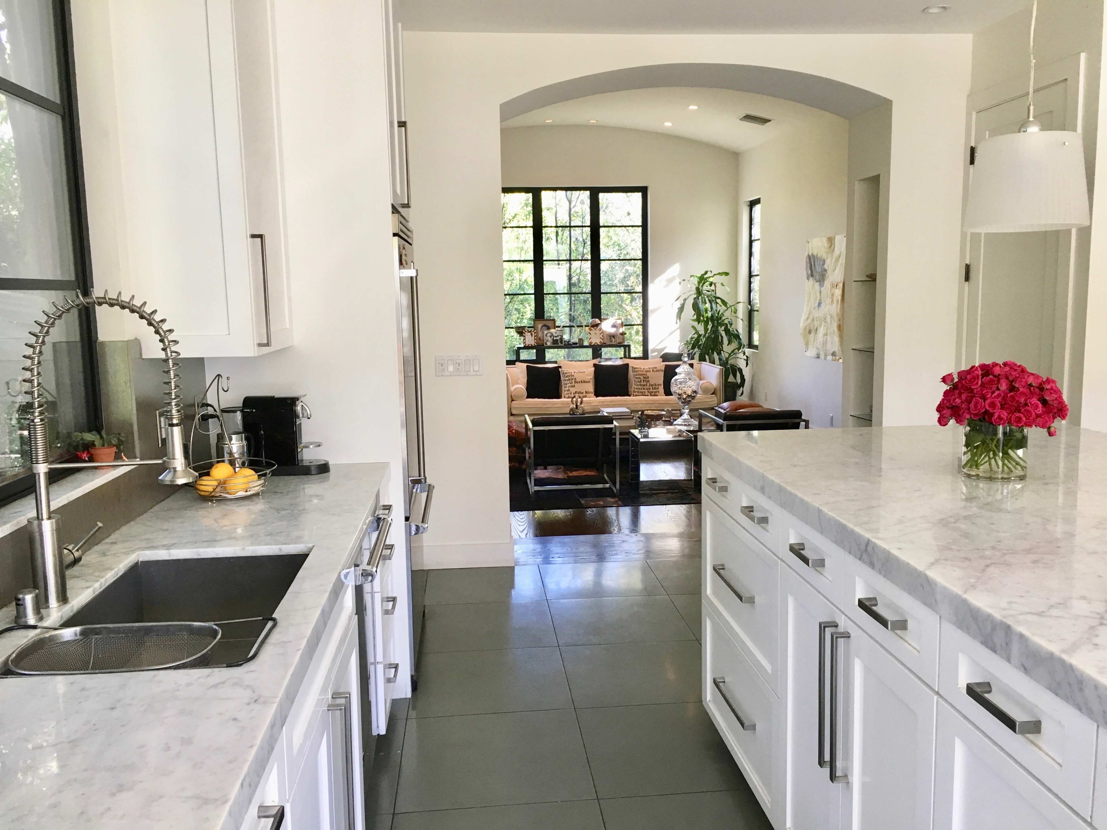 A modern kitchen with white cabinetry and marble countertops opens into a spacious living area visible through an archway.