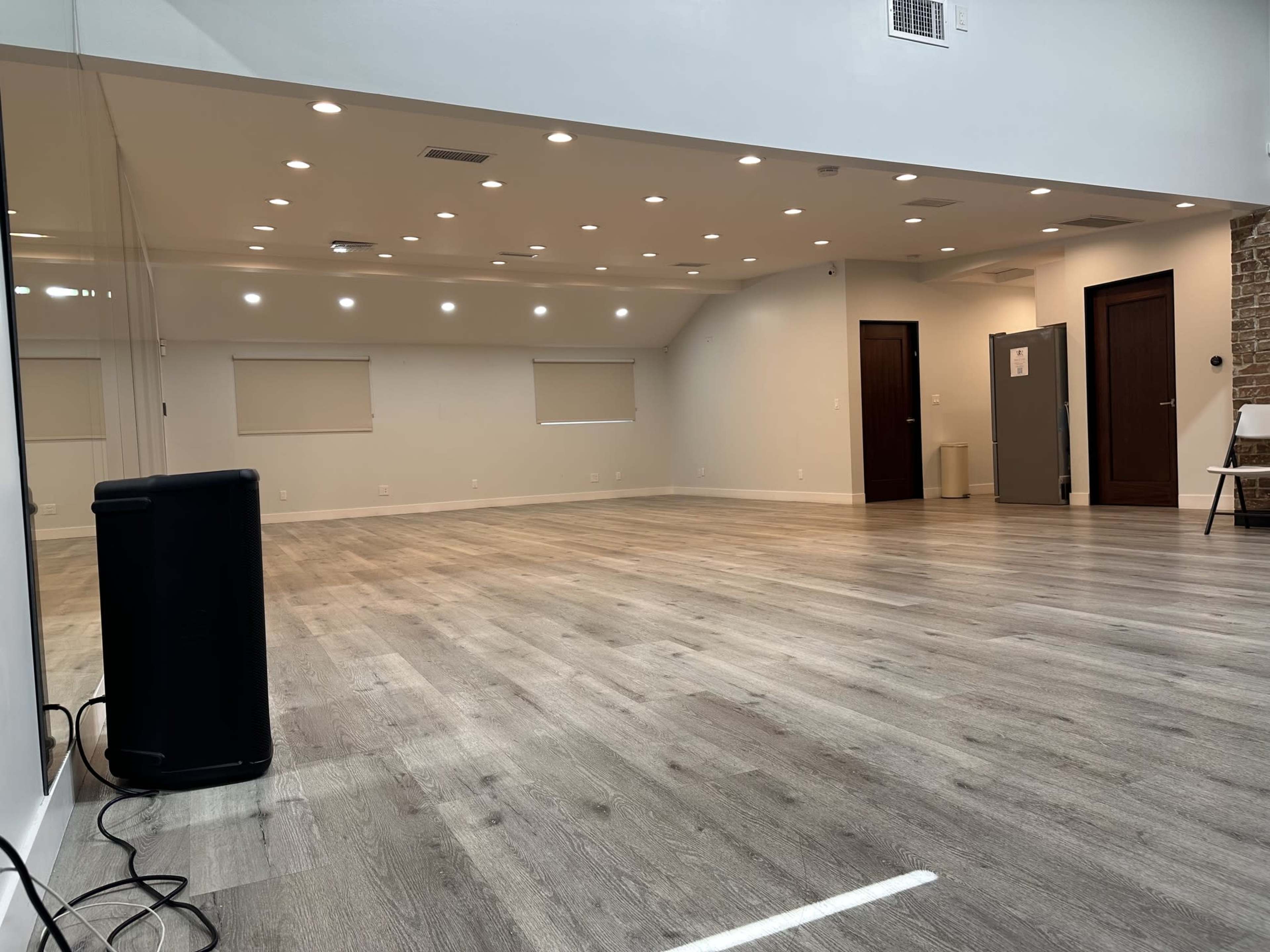 The image shows an empty room with wooden flooring, white walls, and multiple recessed lights, featuring a large mirror on one side and a few doors along the walls.