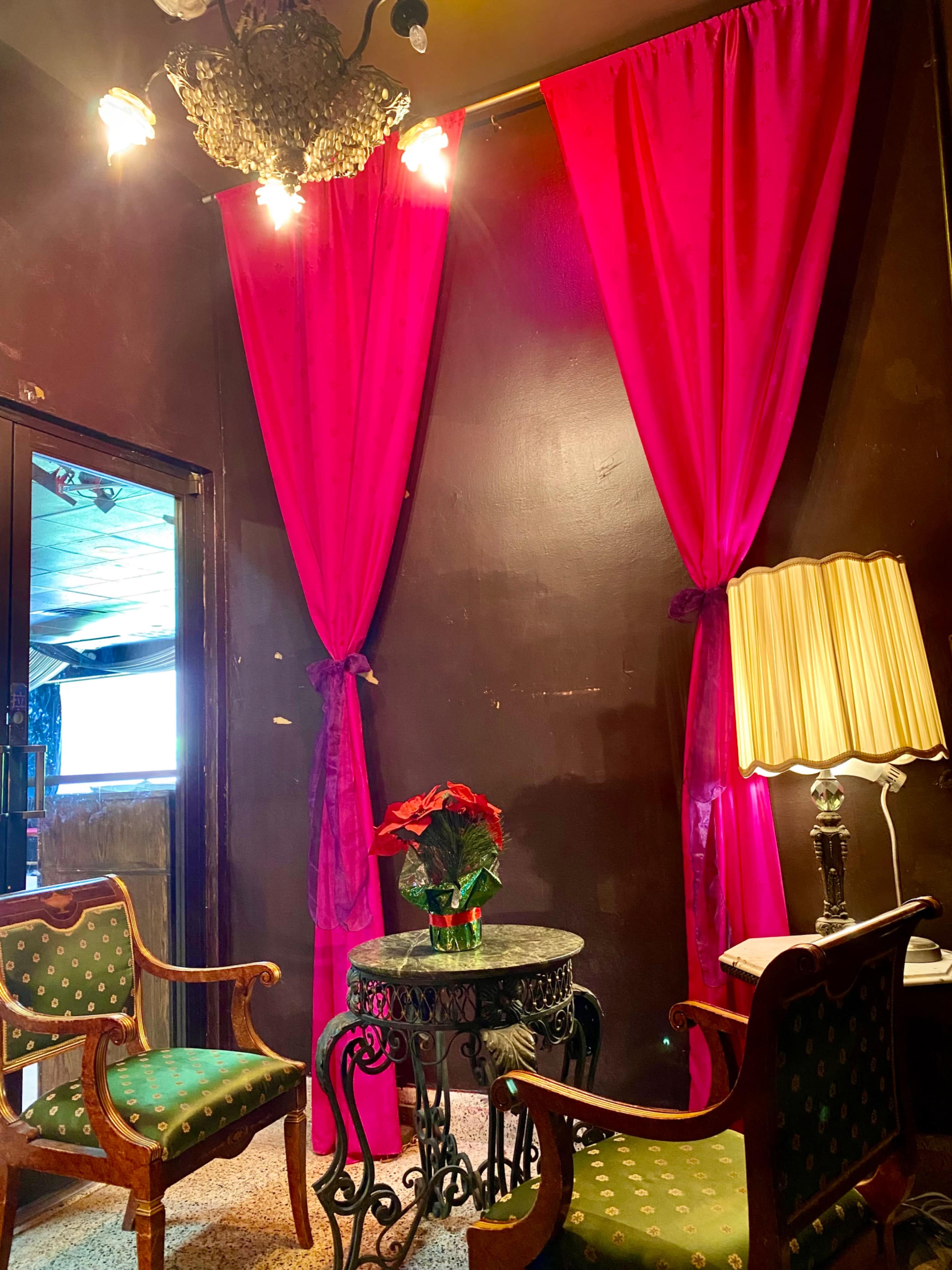 A cozy corner with two chairs, a small table, a lamp, and vibrant pink curtains draping against a dark wall.
