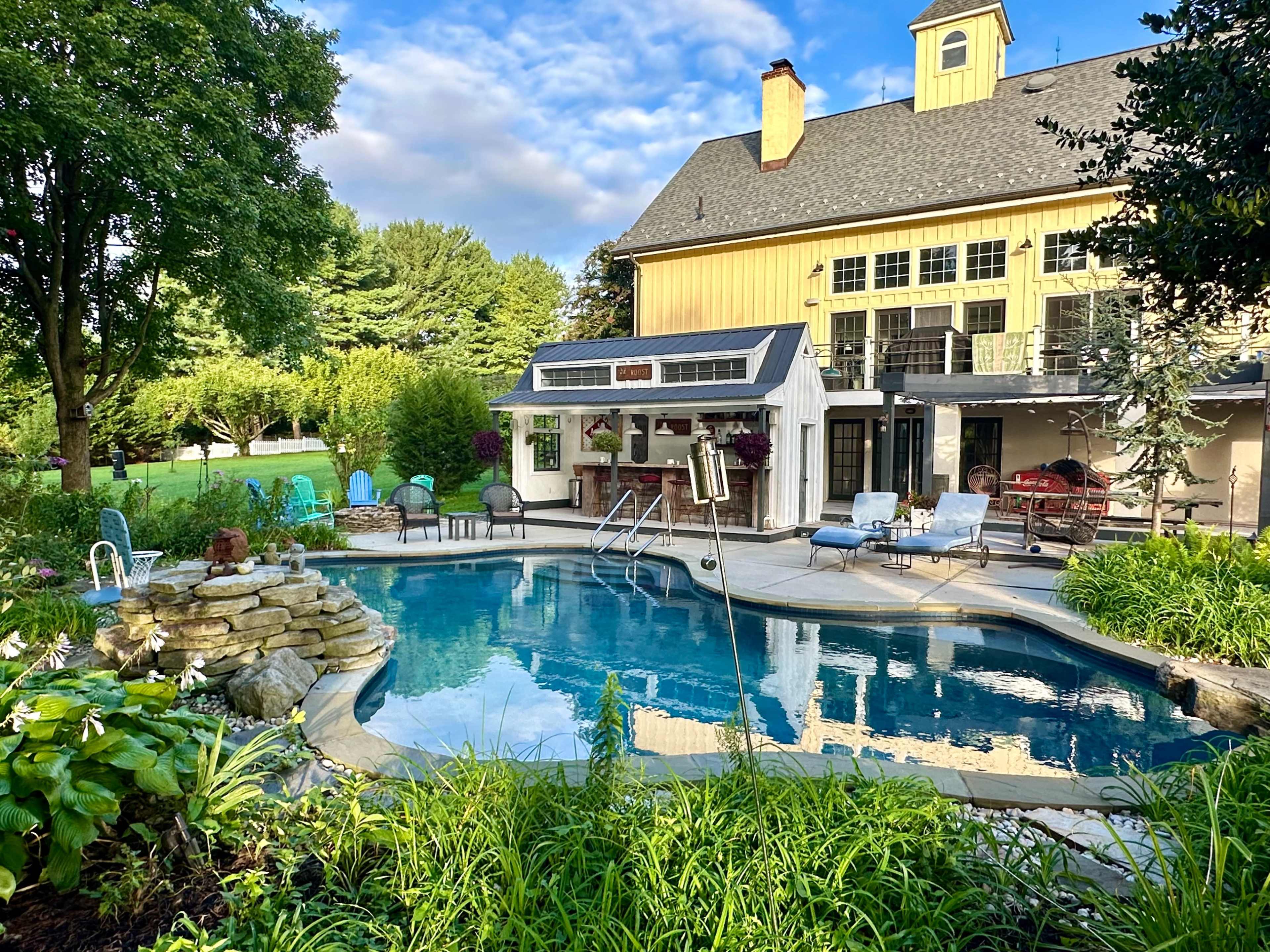 The image shows a backyard pool surrounded by landscaped gardens and a house with a deck in the background.