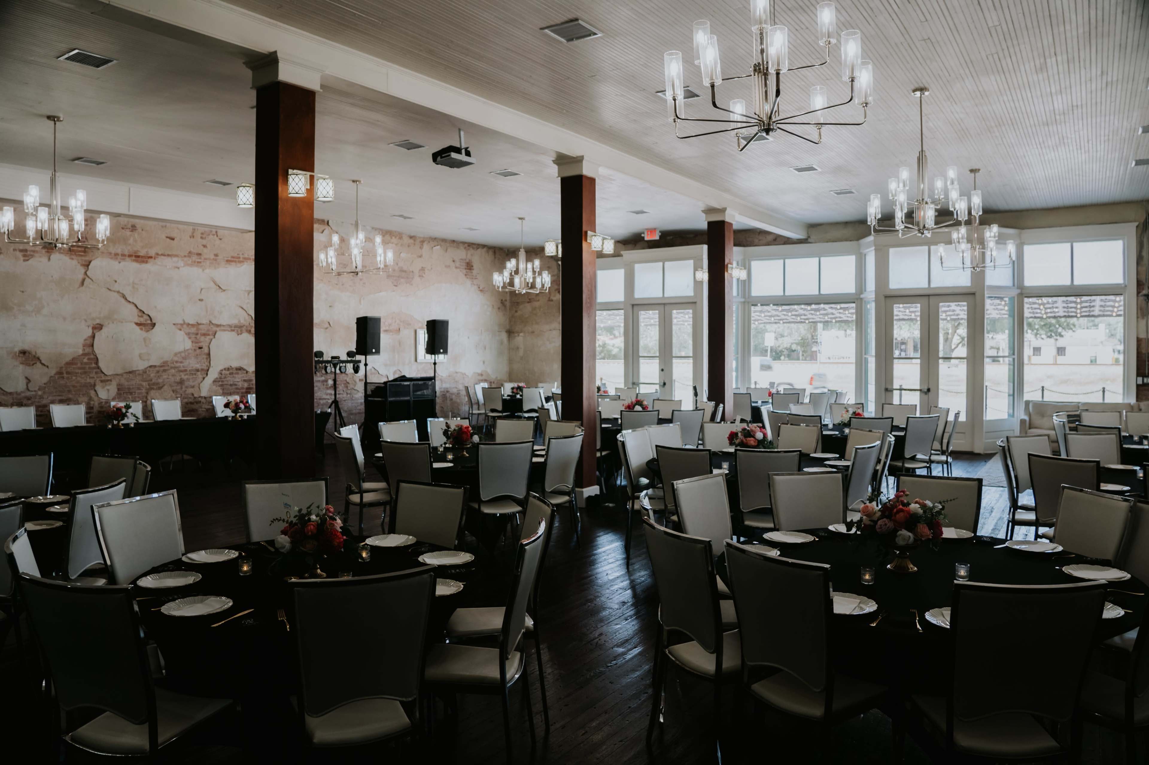 The image shows a spacious event venue with several round tables set with black tablecloths and white chairs, adorned with floral centerpieces, and featuring large windows that provide natural light.