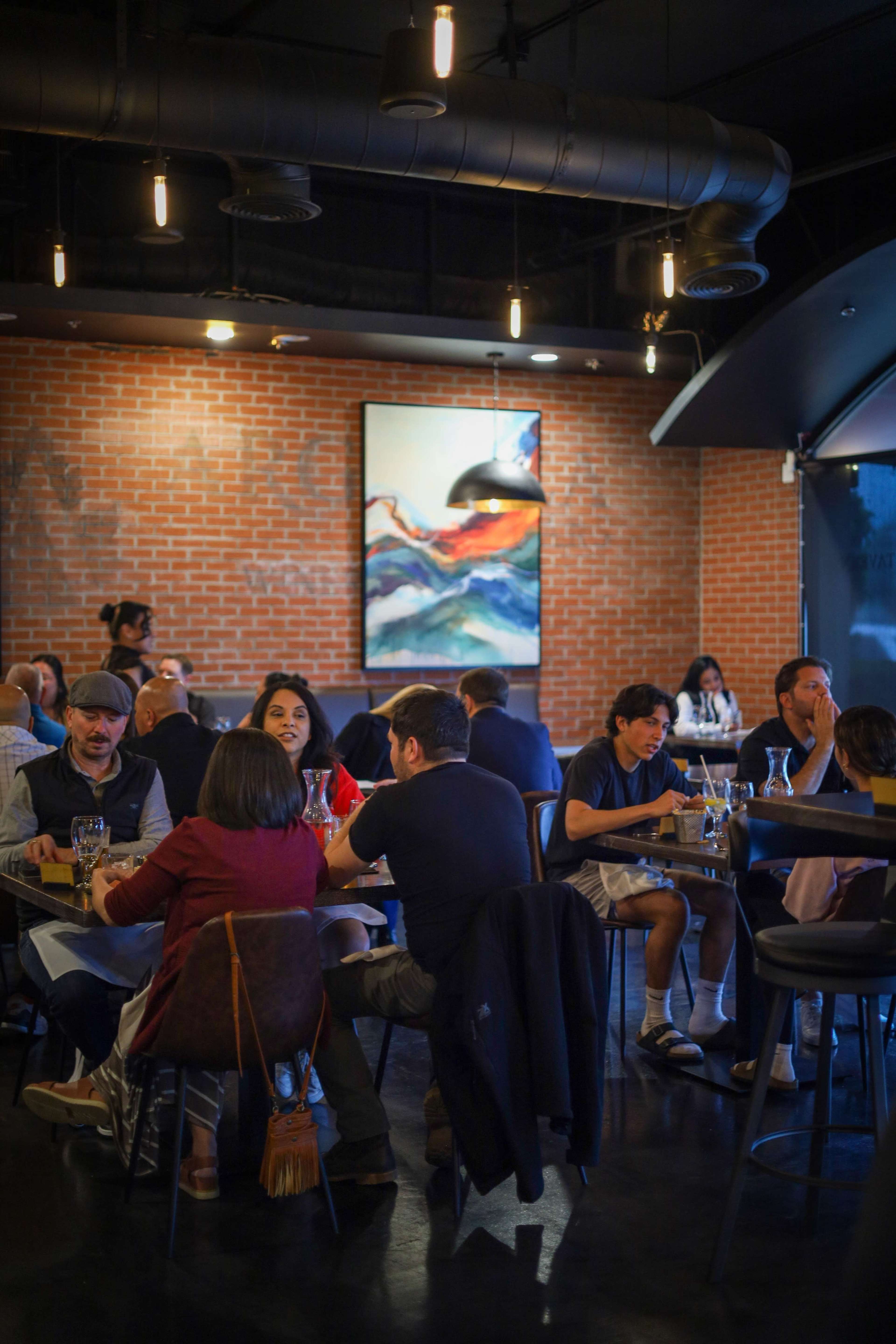 A bustling restaurant interior features patrons dining at tables, with a large, colorful artwork displayed on a brick wall.