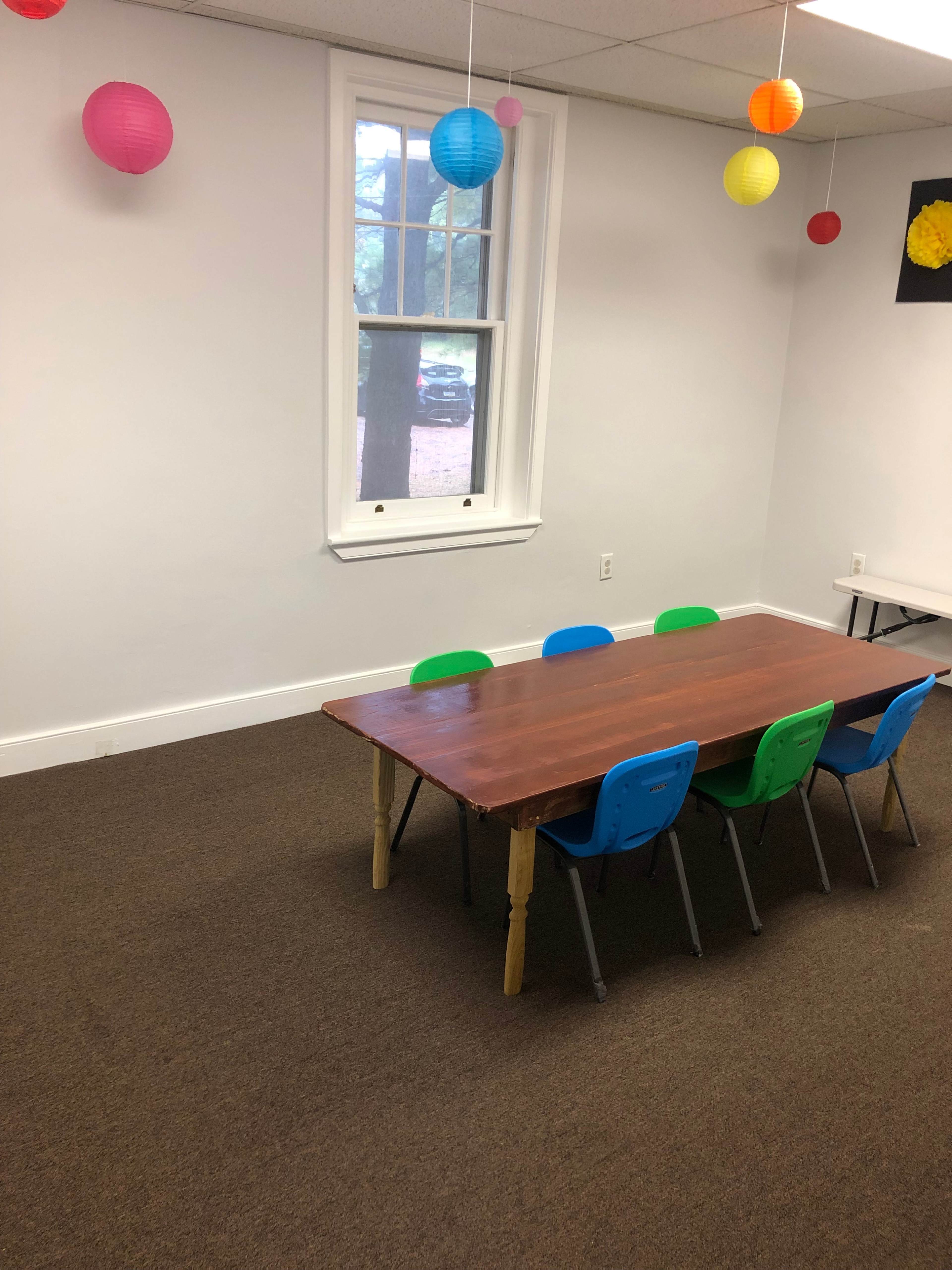 A wooden table surrounded by four colorful chairs is set in a room with a window and paper lanterns hanging from the ceiling.