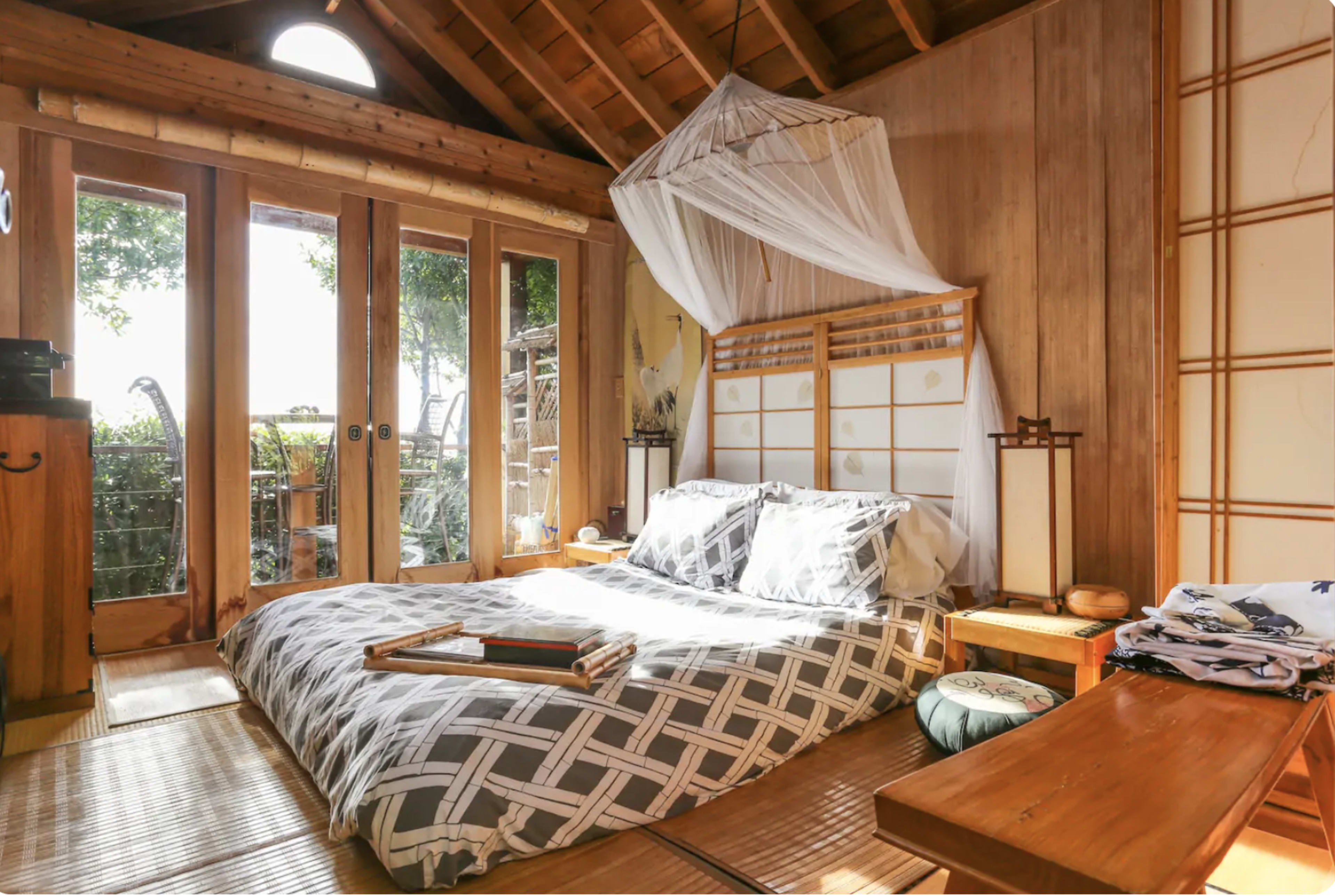 A cozy bedroom features a bed with a patterned duvet, wooden furniture, and large windows opening to a view of greenery.